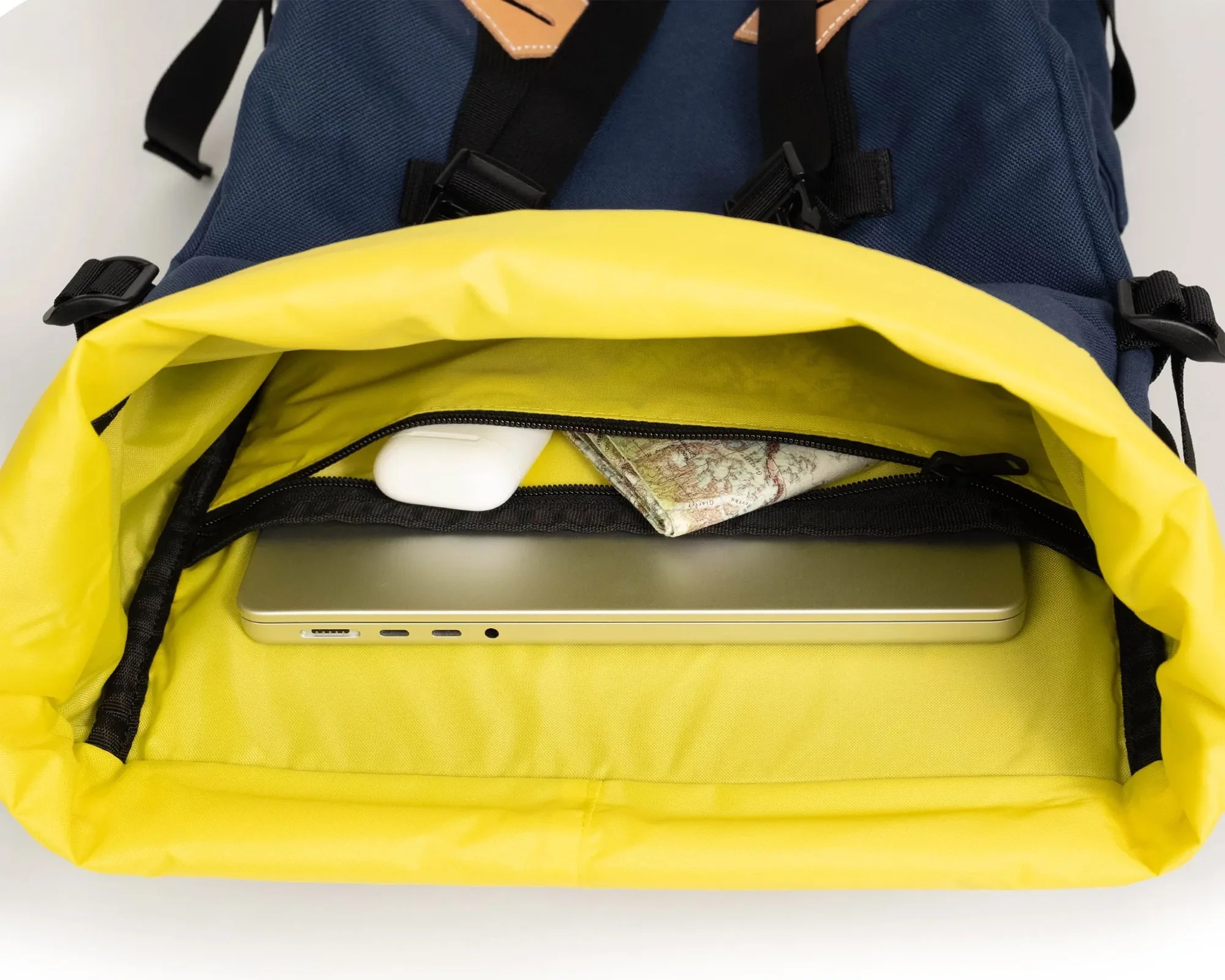 Open navy and yellow backpack showing a silver laptop, white wireless earbuds case, and a folded map inside.