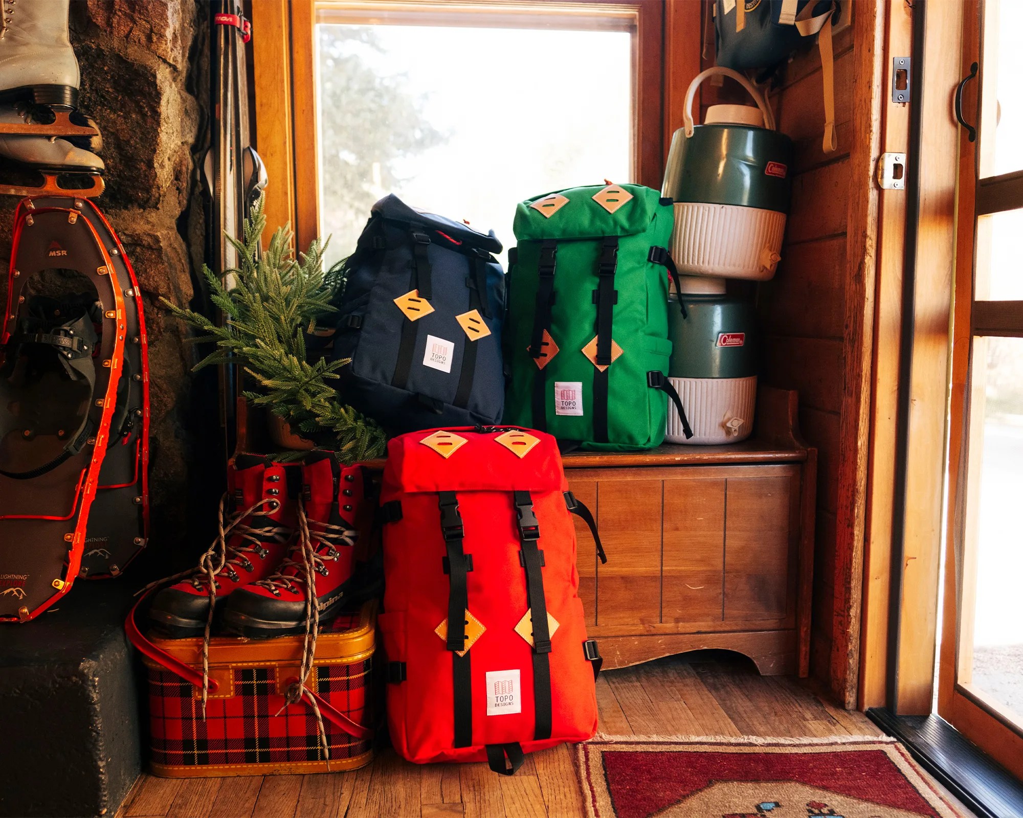 Three colorful Topo Designs backpacks in red, green, and navy blue stacked near a wooden cabinet with Coleman water coolers and red hiking boots on a plaid suitcase.
