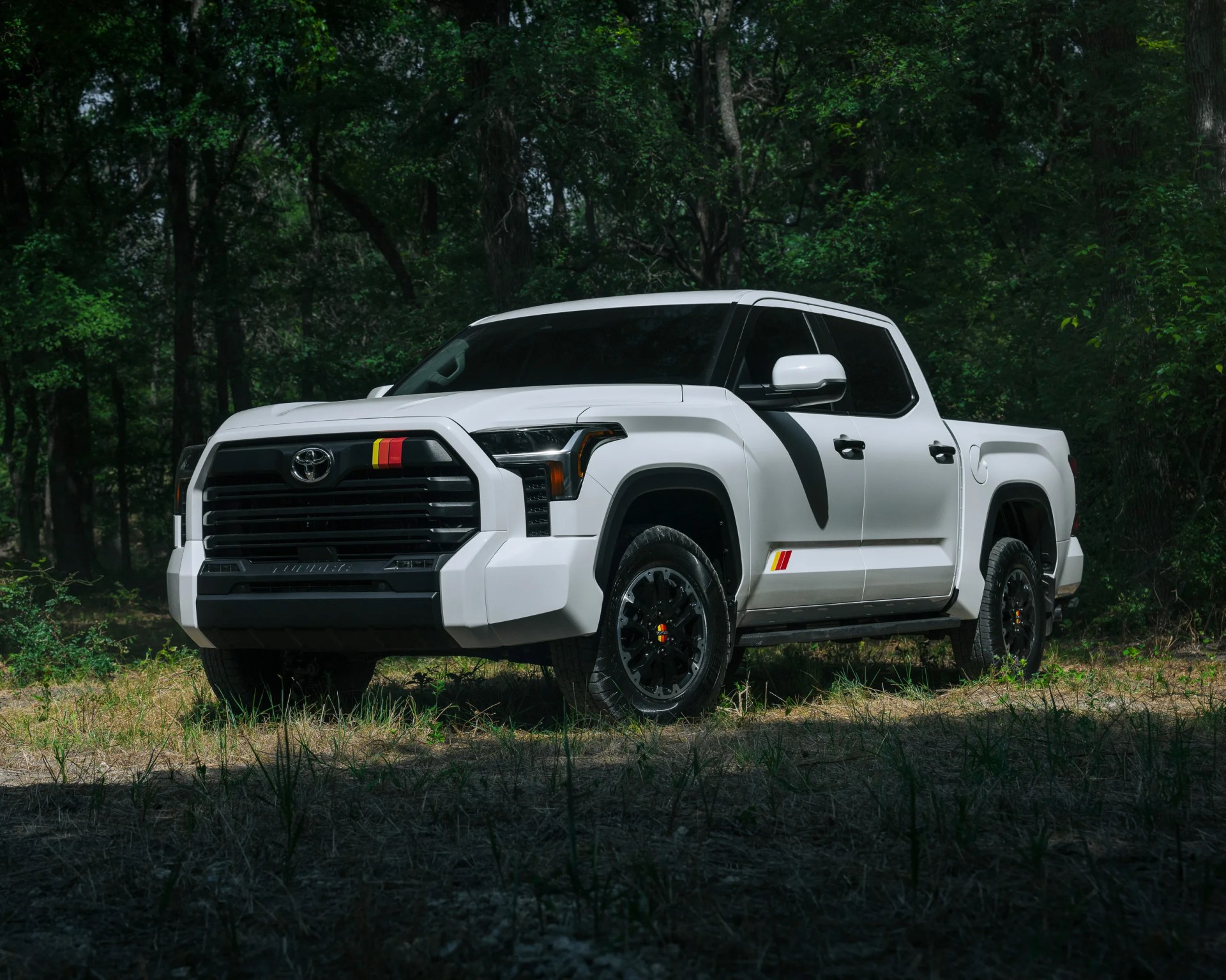 White Toyota pickup truck with black grille and red-yellow stripe decals parked on grass in a forest.