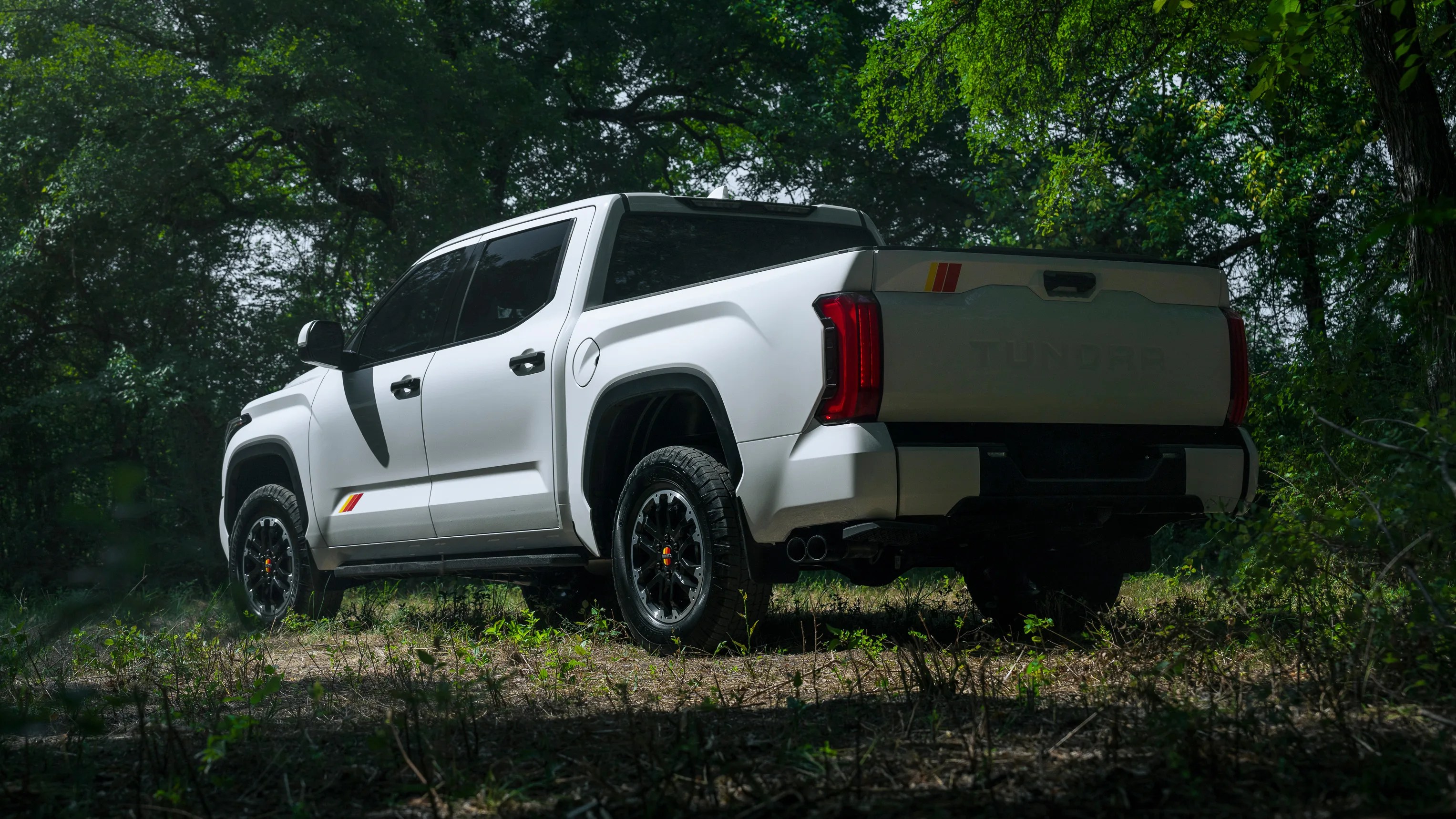 White Toyota Tundra pickup truck with black wheels parked on a dirt path surrounded by green trees.