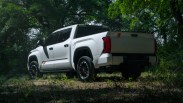 White Toyota Tundra pickup truck with black wheels parked on a dirt path surrounded by green trees.