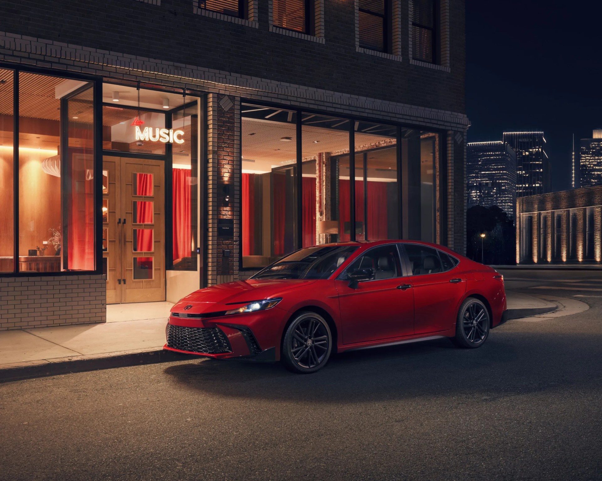 Red sedan parked on a city street at night in front of a building with large windows and a "Live Music" neon sign.