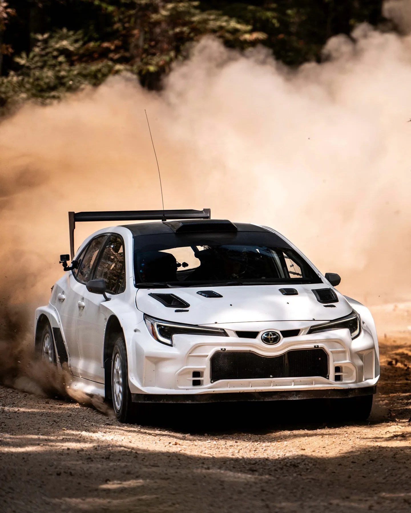 White Toyota rally car kicking up dust on a dirt road with a large rear spoiler.