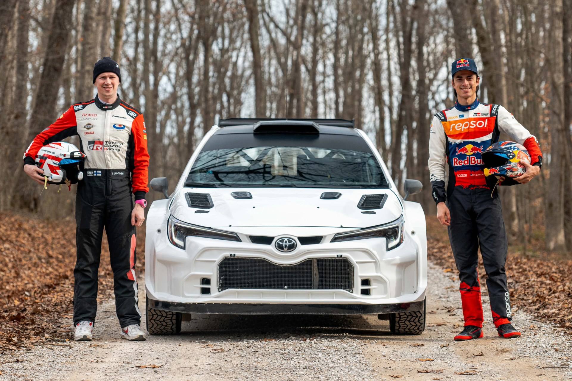 Two race drivers in racing suits holding helmets stand on either side of a white Toyota rally car on a forest dirt road.