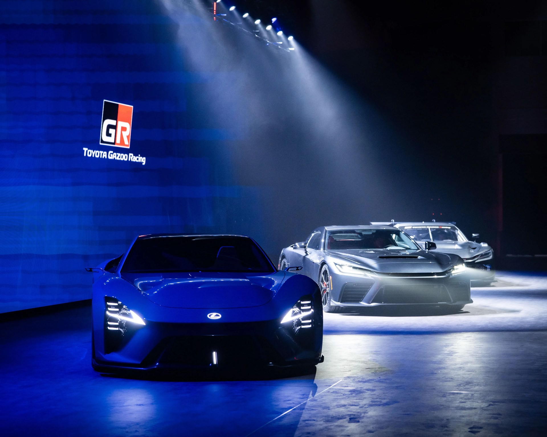 Blue Lexus sports car with headlights on in front of two silver sports cars under spotlights in a dark showroom with Toyota Gazoo Racing logo on the wall.
