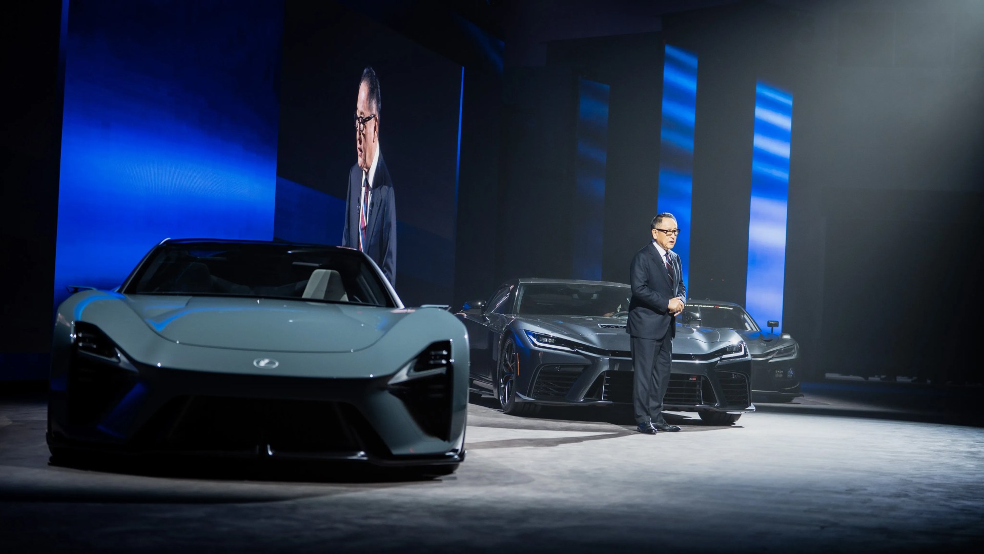 Man in a suit standing next to two dark gray sports cars on a stage with blue vertical light panels.