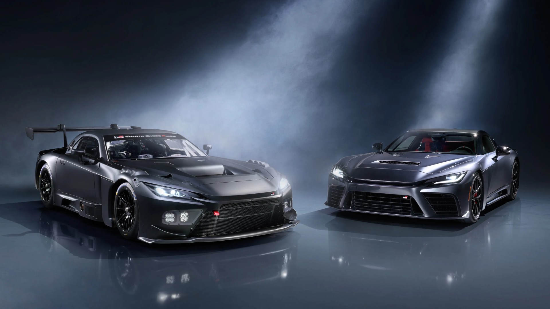 Two dark gray Toyota GR sports cars with headlights on, one race-modified with a large rear wing and one street version, in a smoky studio.