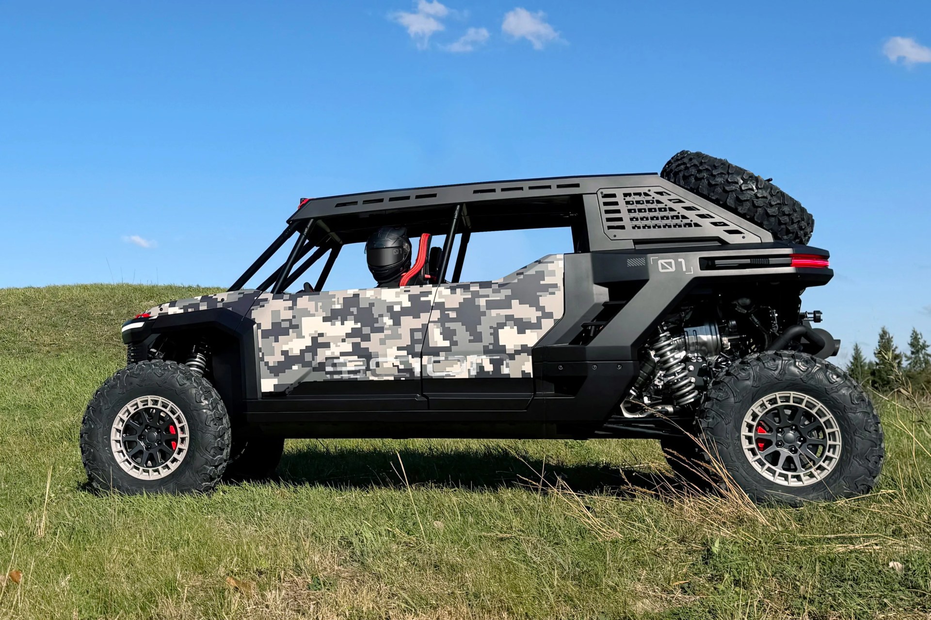 Off-road vehicle with digital camouflage pattern and large rugged tires on grass under blue sky.