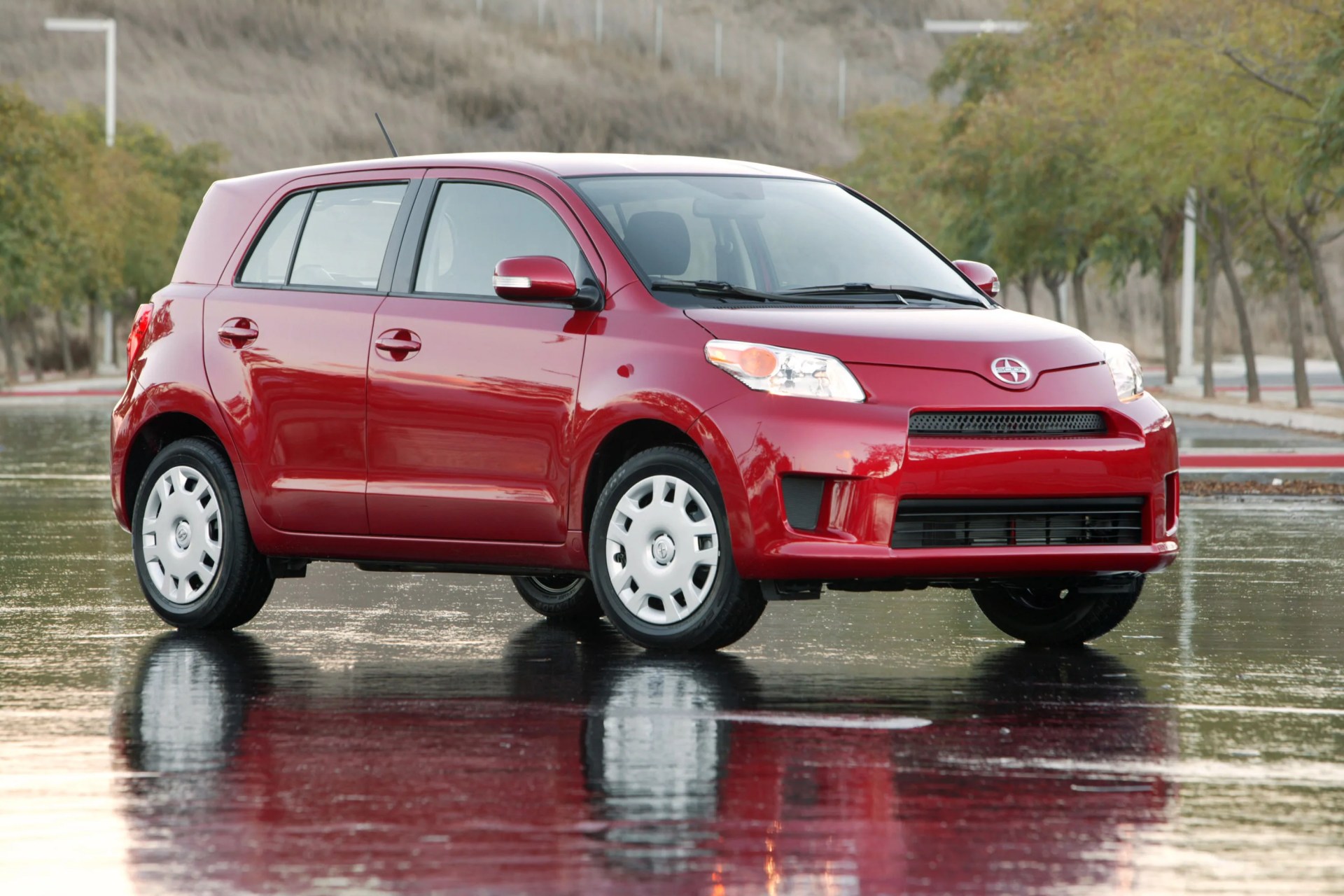 Red compact hatchback car parked on a wet reflective surface with trees in the background.
