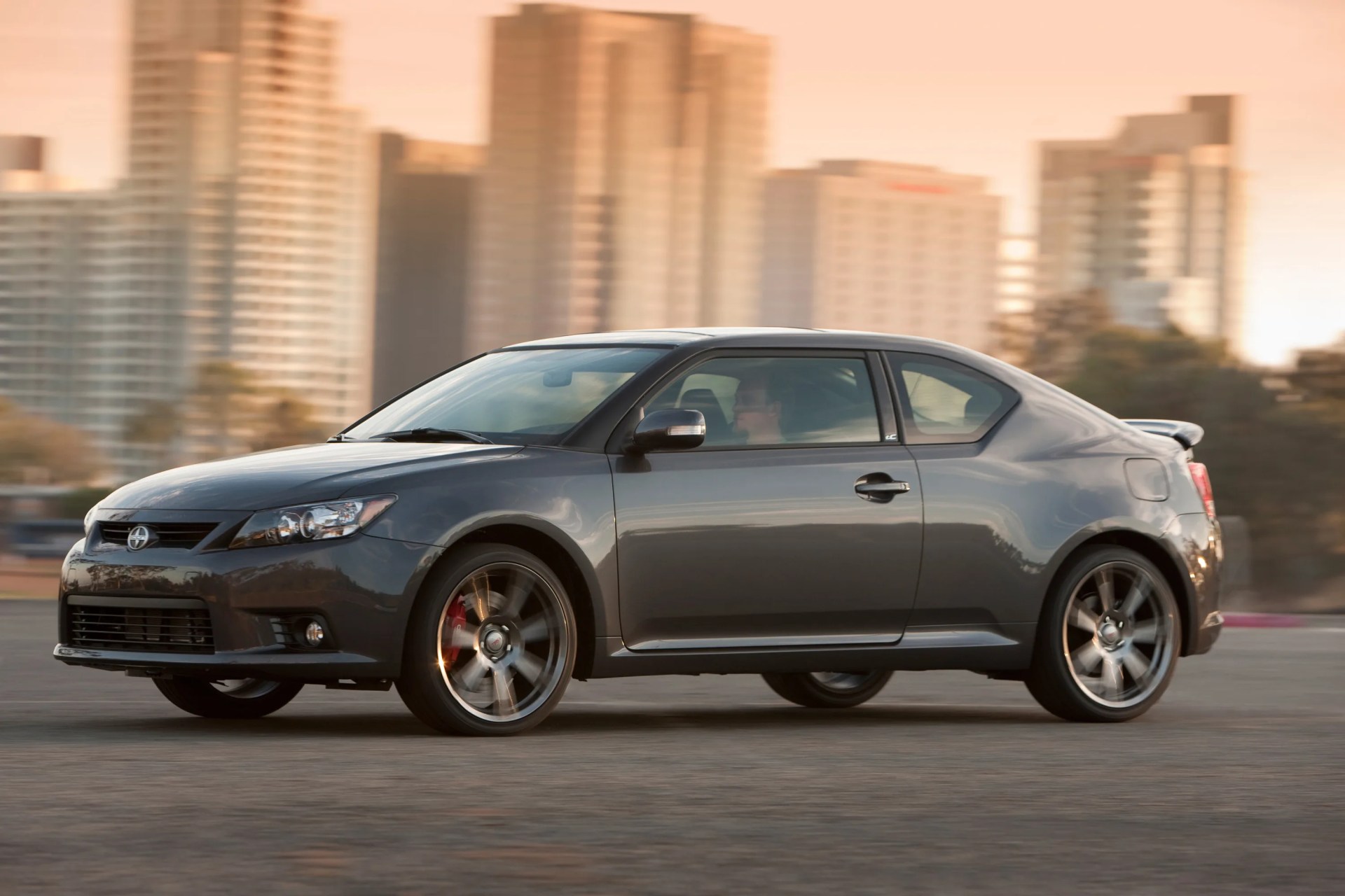 Gray Scion tC coupe with alloy wheels driving on a city street at sunset.