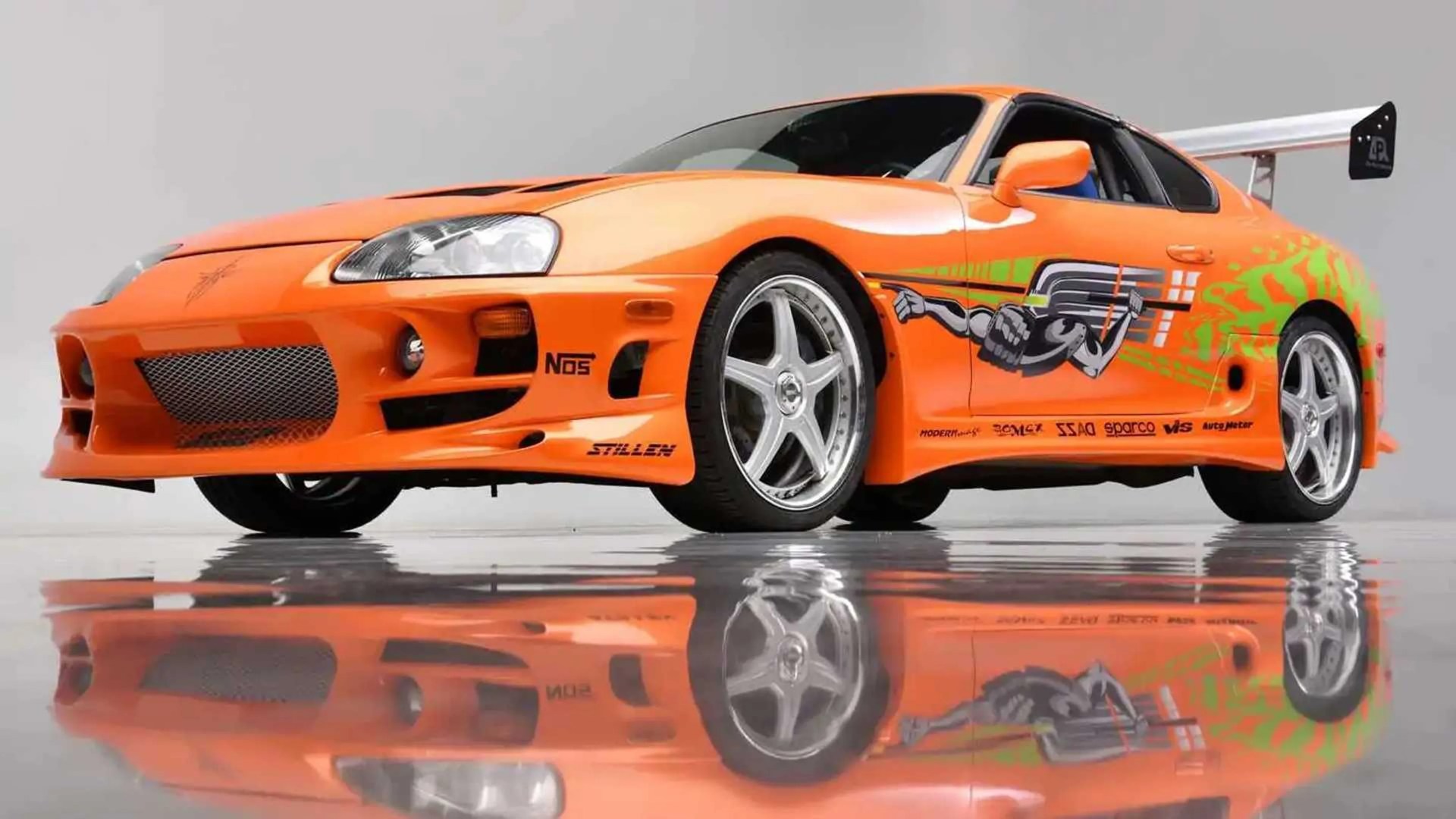 Orange sports car with racing decals, silver five-spoke wheels, and a large rear spoiler on a reflective floor.