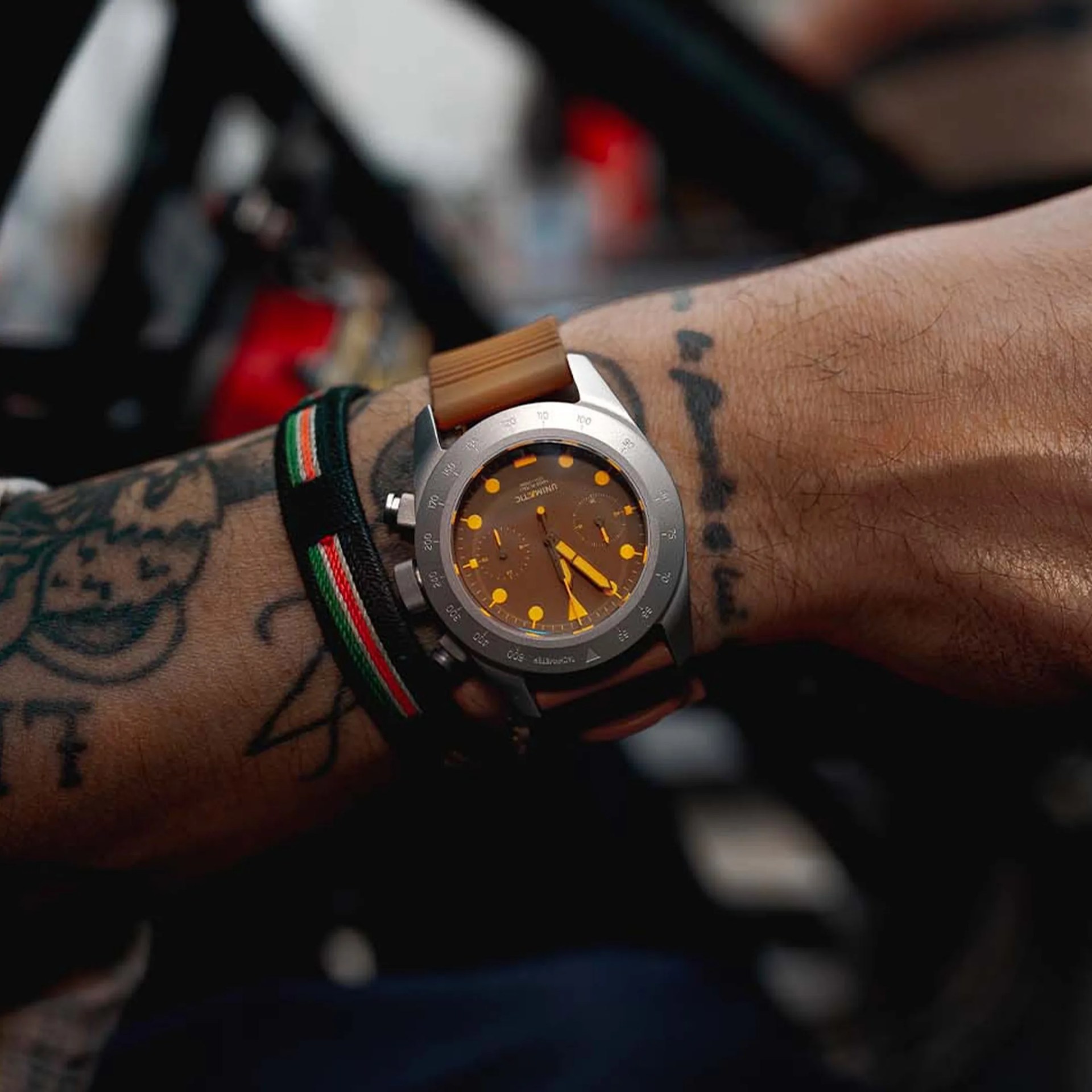 Wristwatch with a brown dial and orange markers on a tan strap worn on a tattooed wrist with a black, green, white, and red striped bracelet.