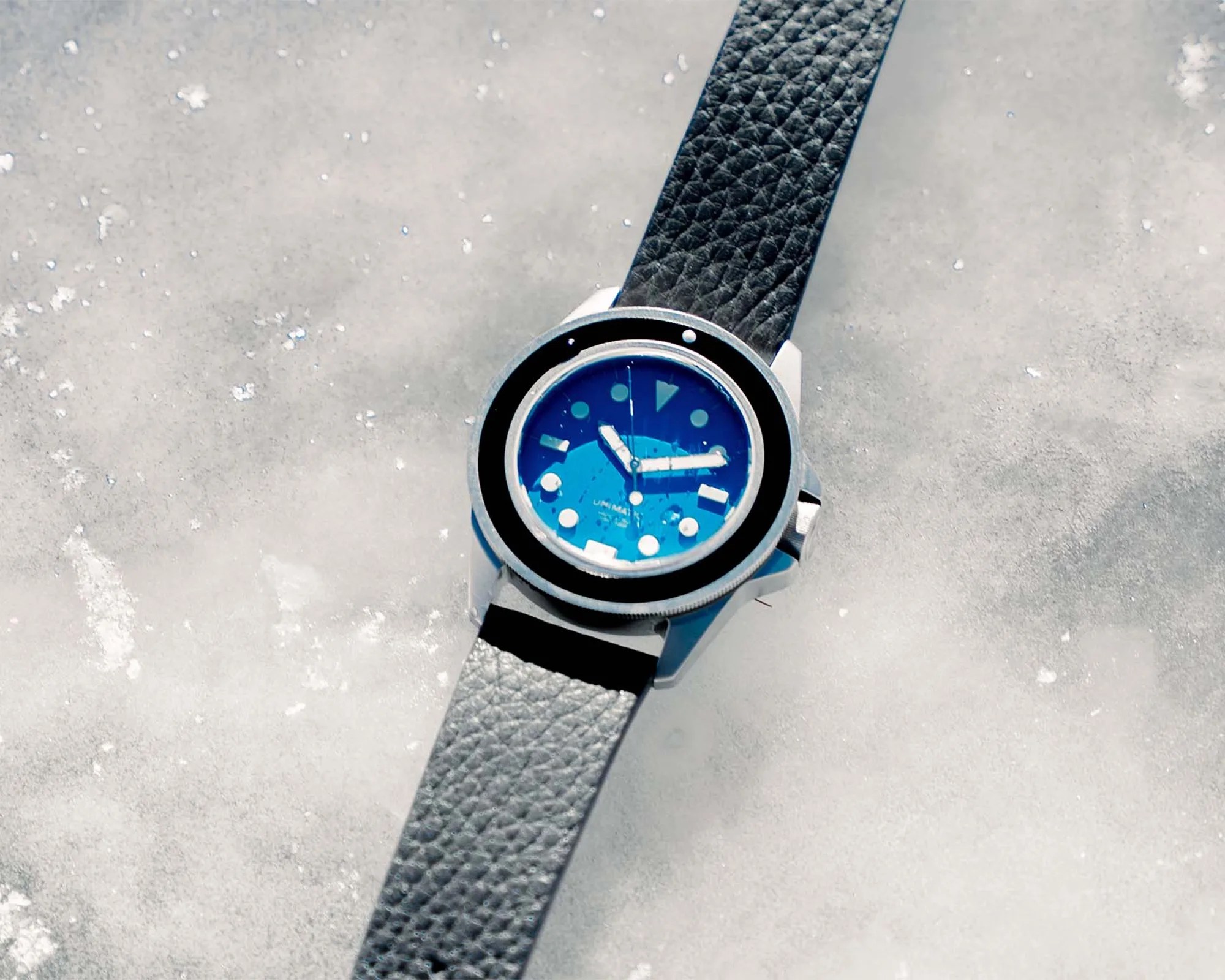 Diver-style wristwatch with a blue dial, black bezel, and black textured leather strap on a snowy surface.