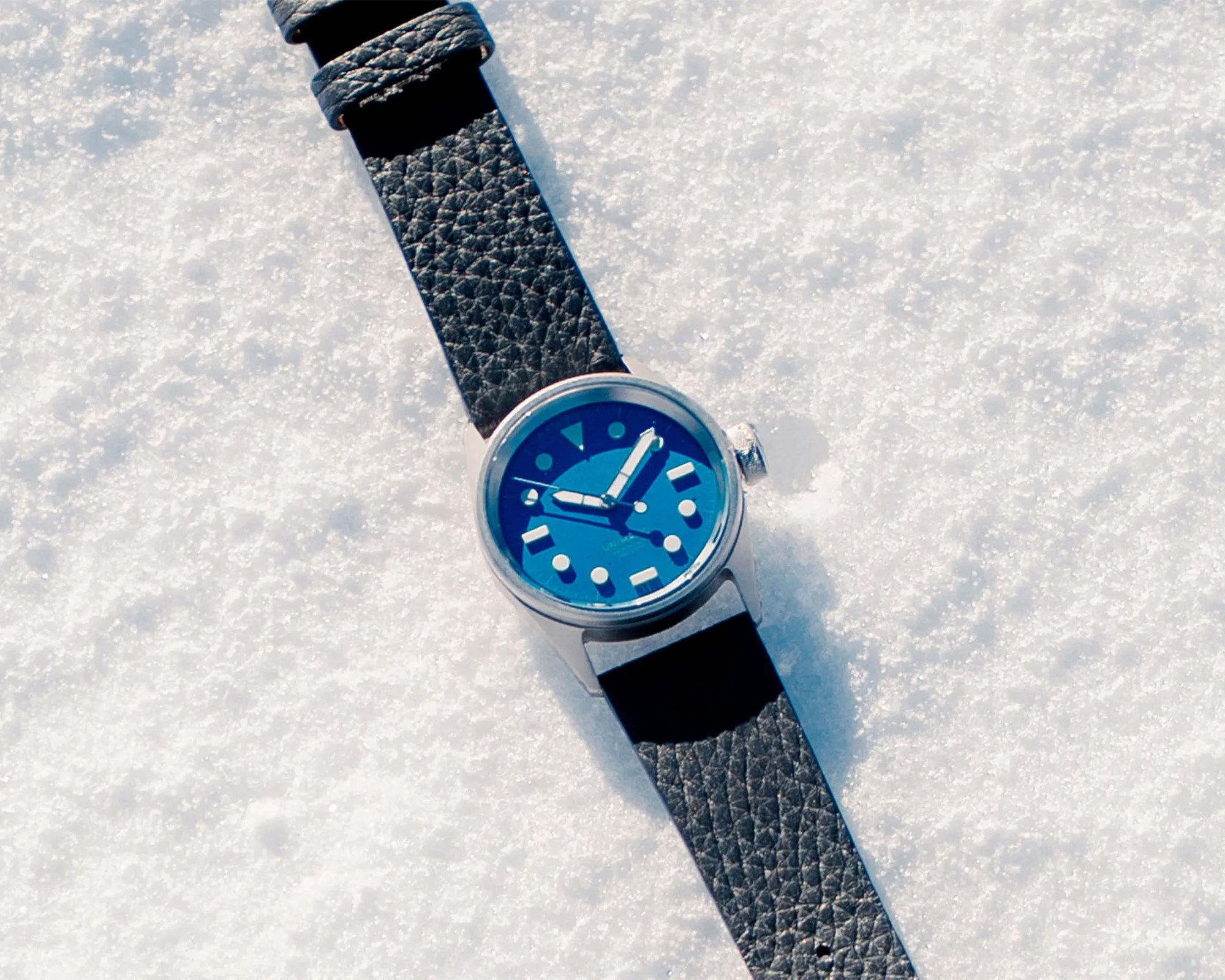 Silver wristwatch with a blue dial and black textured leather strap lying on snow.