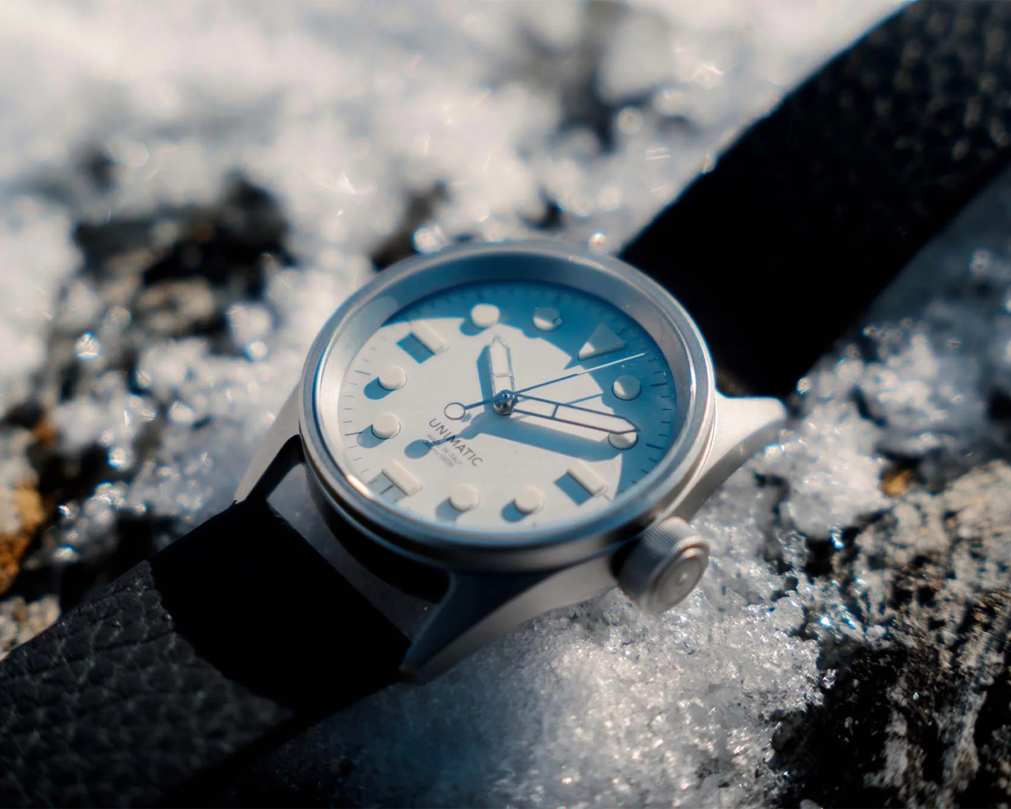 Silver Unimatic wristwatch with white dial and black leather strap on snow-covered ground.