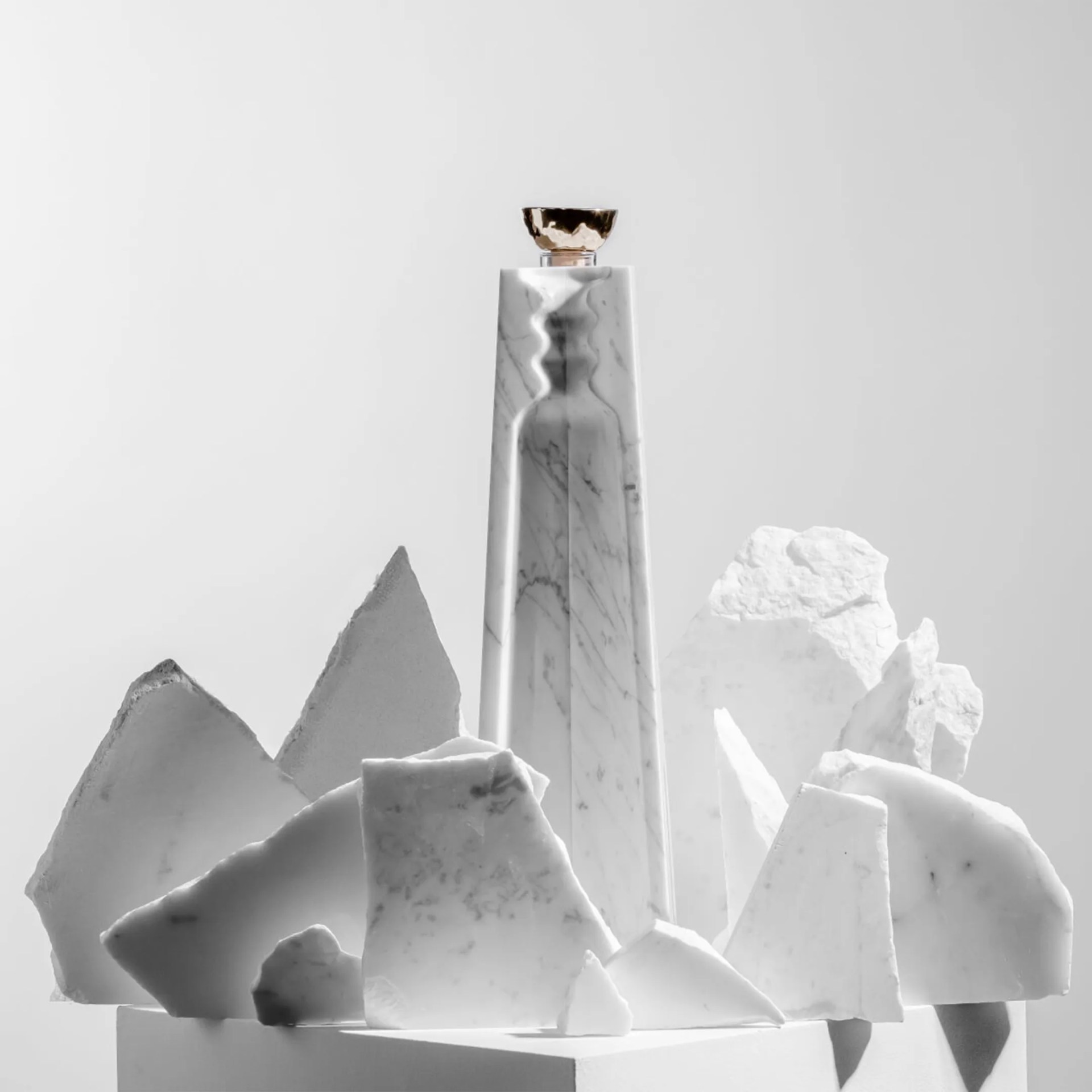 Tall white marble bottle with a gold cap surrounded by irregular white marble shards on a white pedestal.