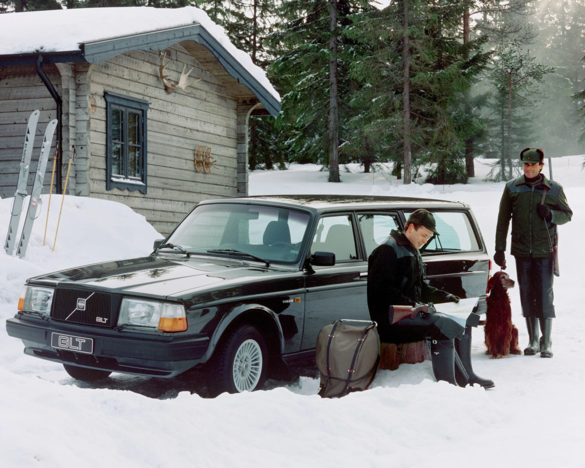 Black Volvo GLT station wagon parked in snow near a wooden cabin with two men in hunting attire and a dog.