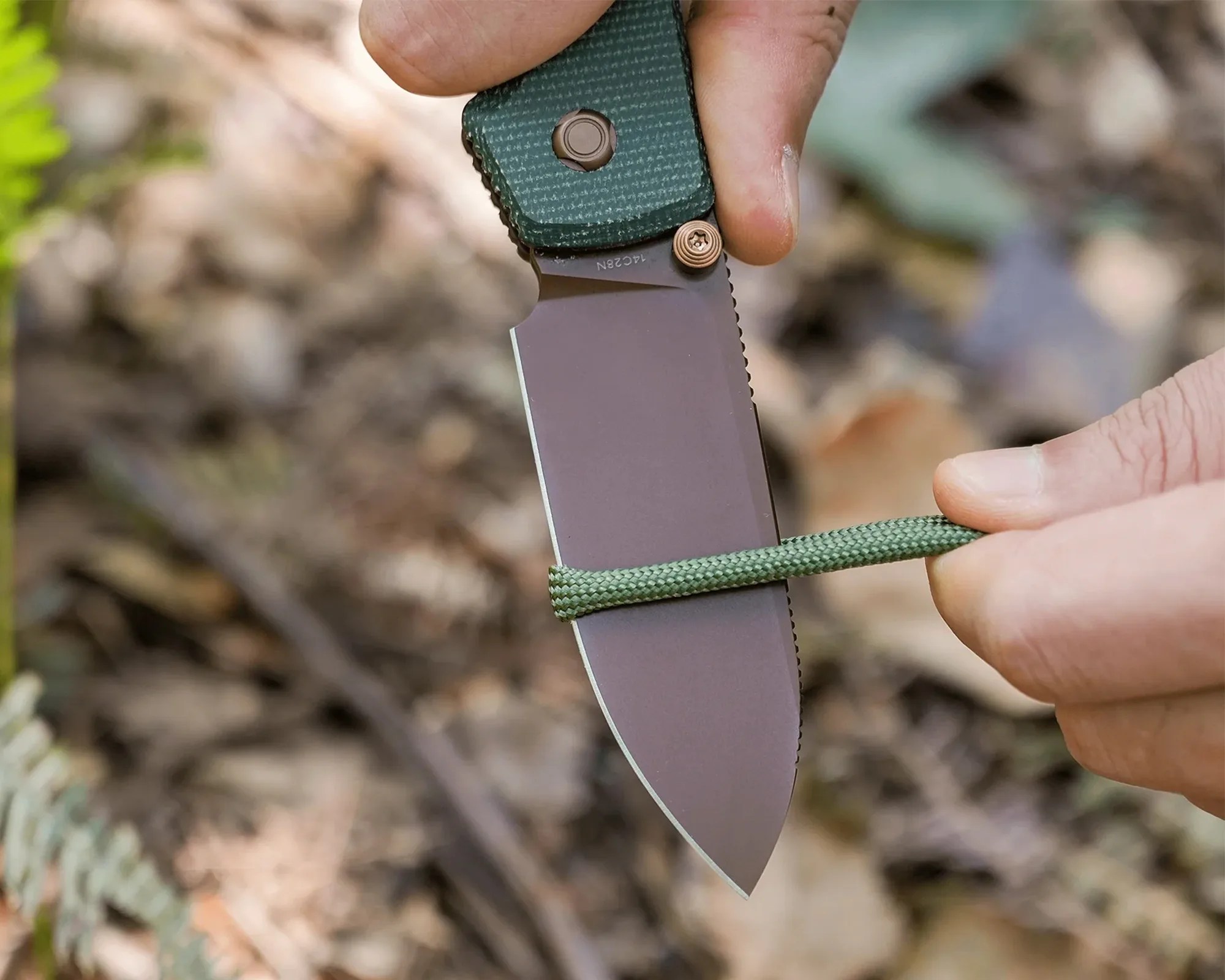 Close-up of a hand holding a green-handled folding knife cutting a green paracord rope outdoors.