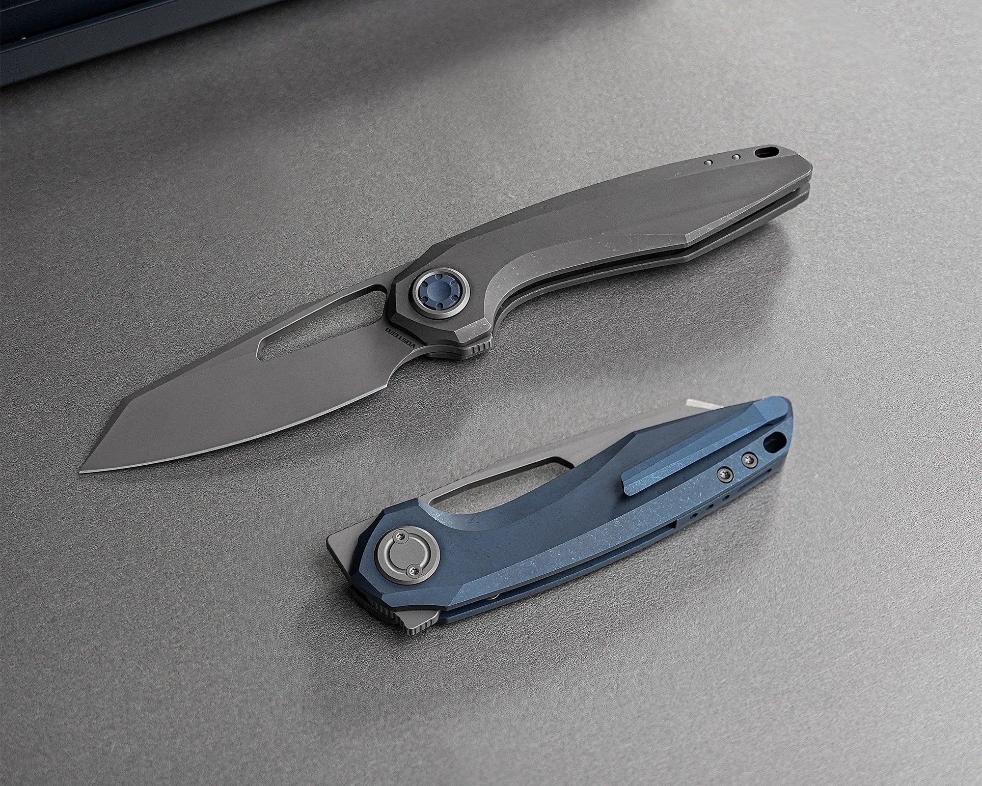 Two folding knives on a gray surface, one open with a black handle and one closed with a blue handle.