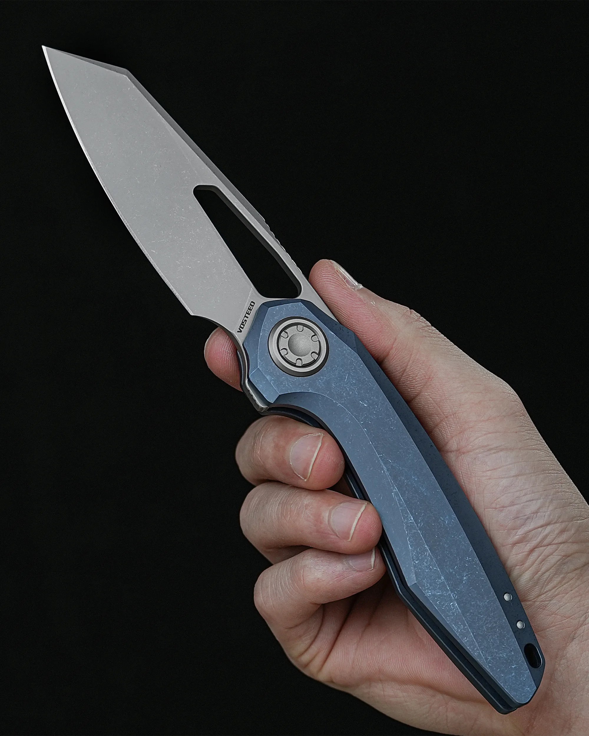 Hand holding a folding knife with a stonewashed blade and blue textured handle against a black background.