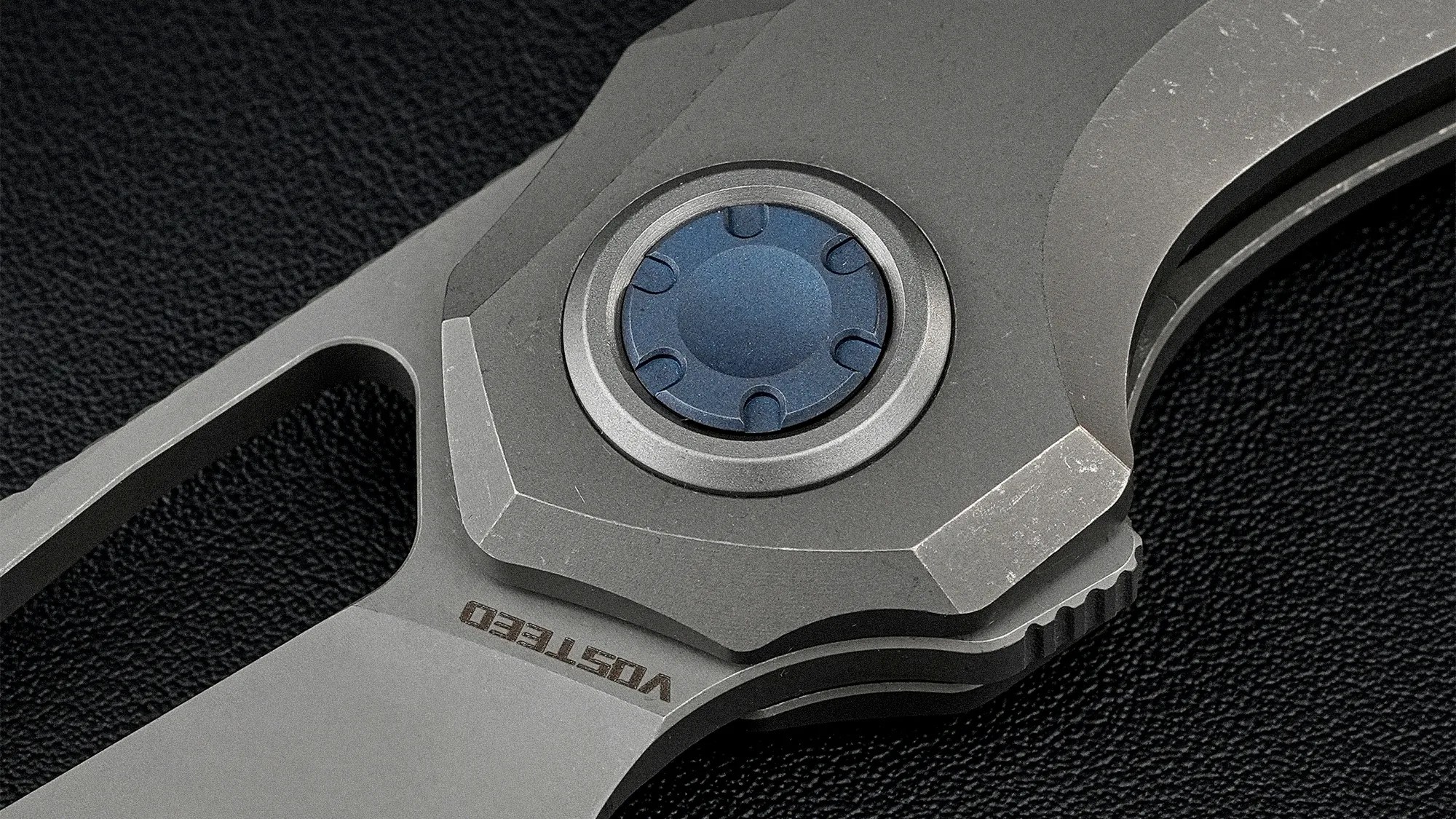 Close-up of a gray folding knife pivot with a blue circular screw and "VOSTED" engraved on the blade.