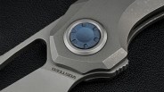 Close-up of a gray folding knife pivot with a blue circular screw and "VOSTED" engraved on the blade.