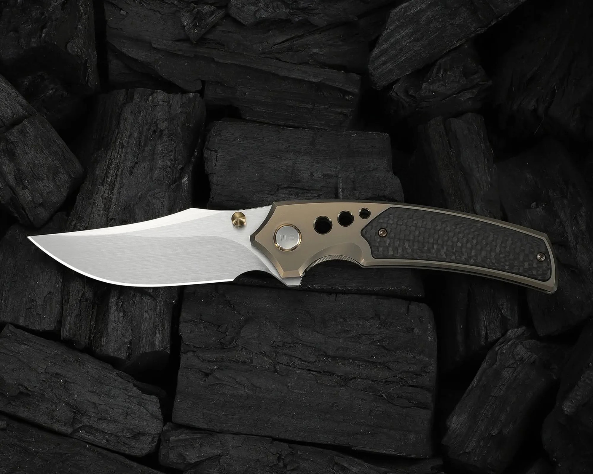 Folding knife with a silver blade, bronze frame, and black textured handle resting on black charcoal pieces.