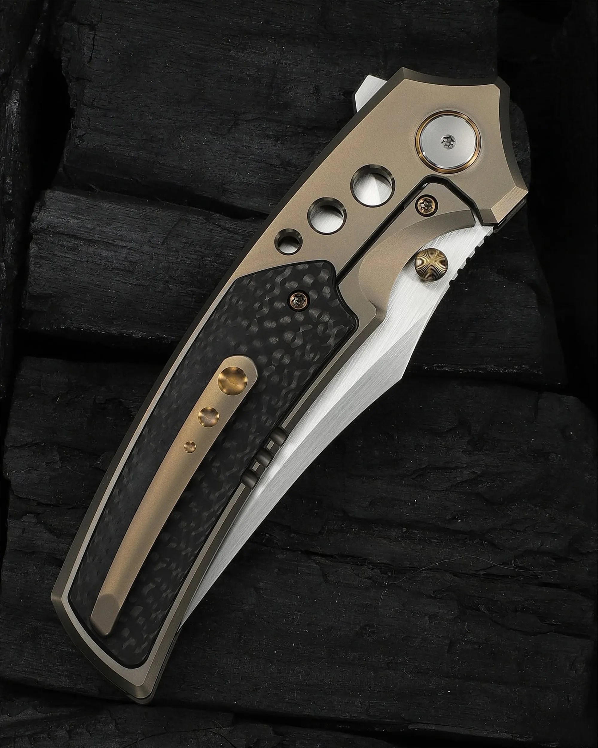 Folding pocket knife with a titanium handle, carbon fiber inlay, and a partially visible stainless steel blade on black textured surface.