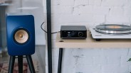 Blue KEF LS50 Meta speaker on a black tripod stand next to a wooden table with a black audio device and white turntable.
