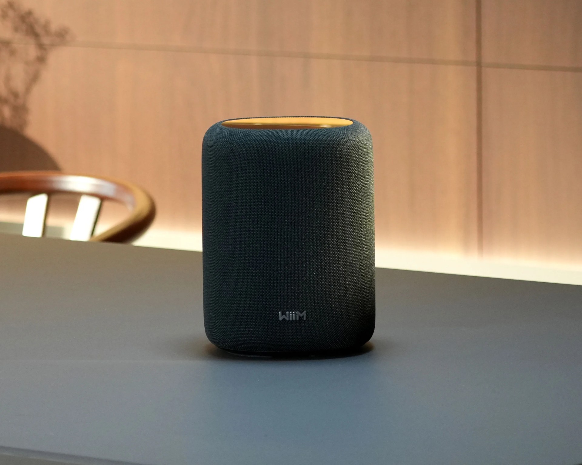 Black cylindrical smart speaker with fabric cover and yellow top on a gray table.