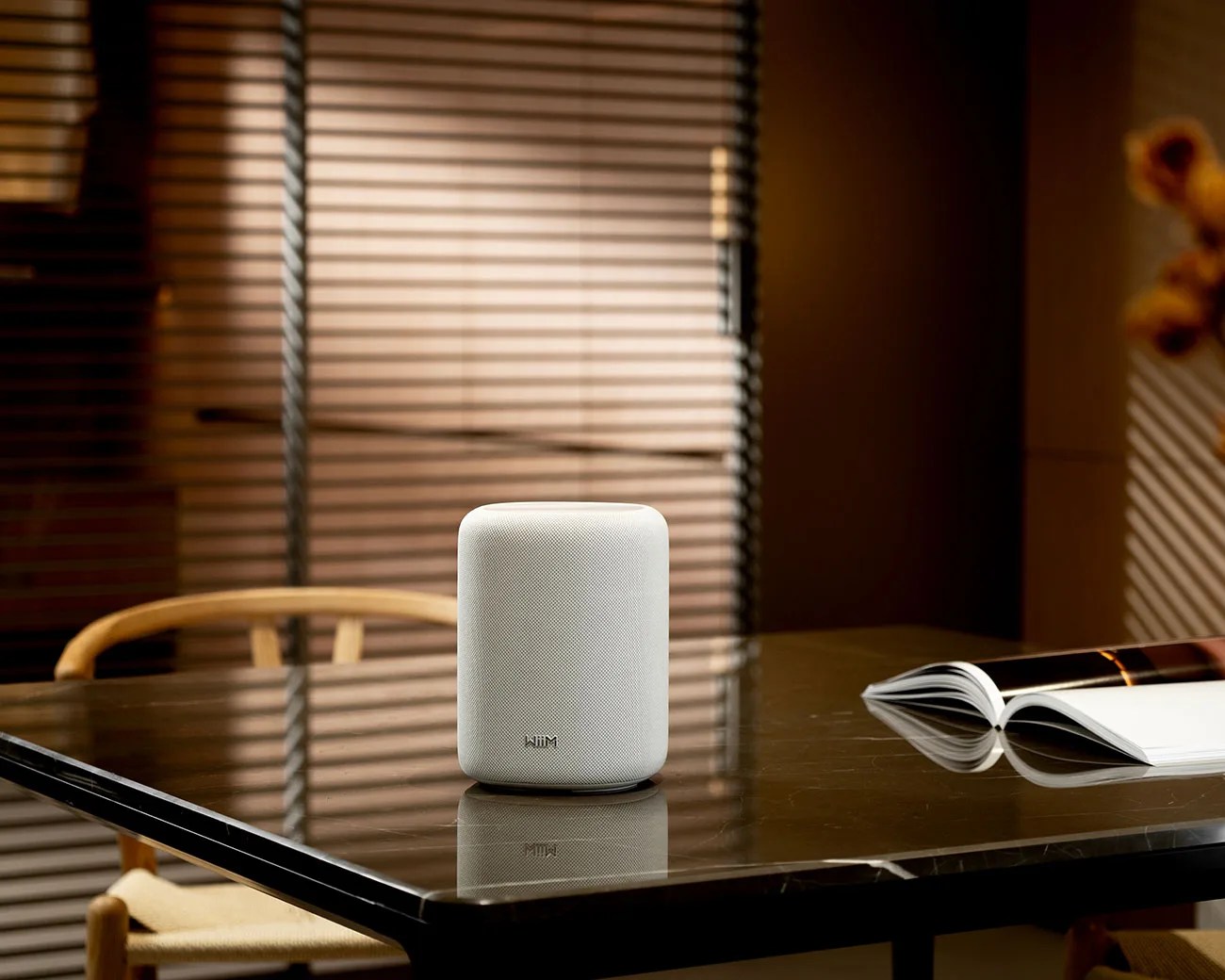 White cylindrical smart speaker with fabric cover on a reflective dark marble table next to an open book.