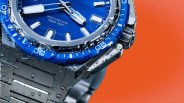 Close-up of a dive watch with a blue bezel and dial, water droplets on the surface, and an orange background.