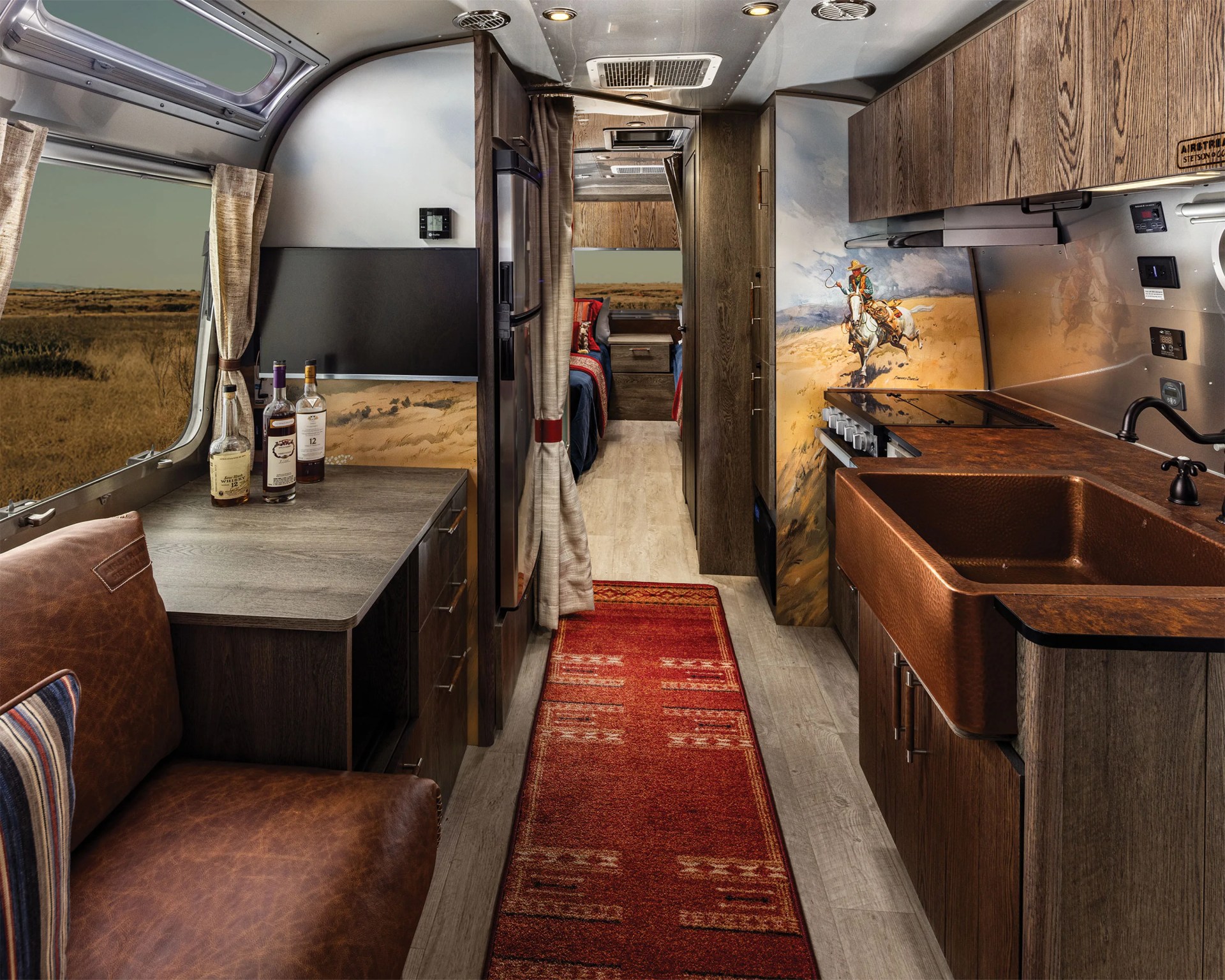 Interior of an Airstream trailer featuring a brown leather couch, wooden cabinets, a copper sink, a stove, and a red patterned rug.