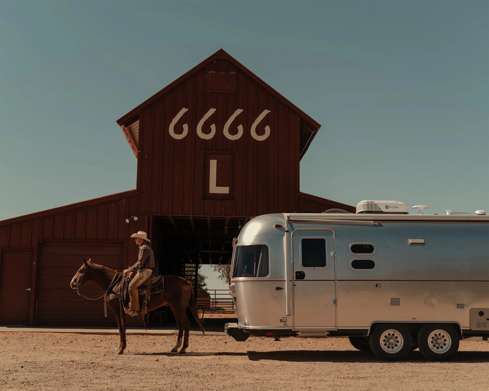 Brown barn with "6666 L" painted on it, a person on a horse, and a silver trailer parked nearby on dirt ground.