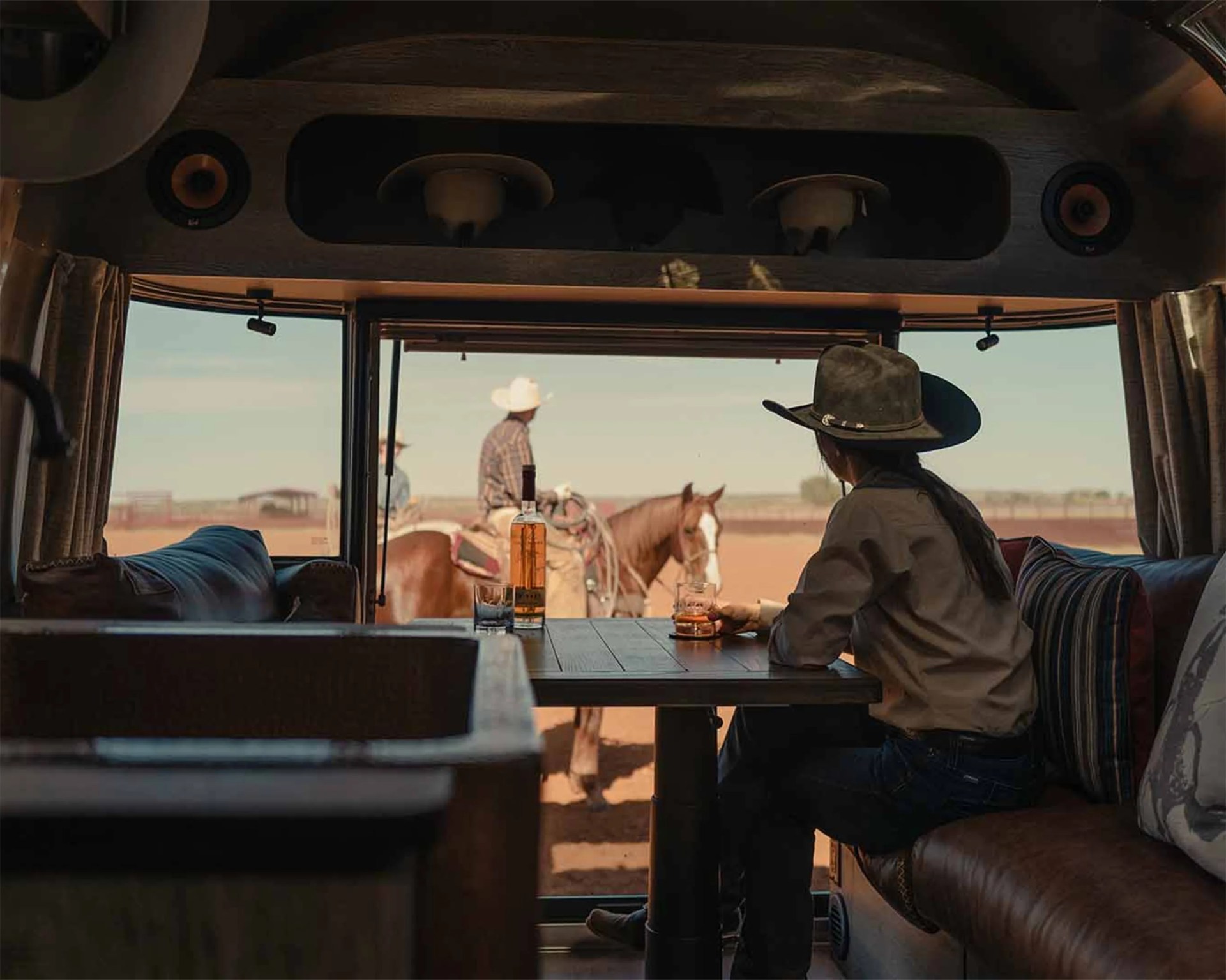 Person wearing a black cowboy hat sitting inside a vehicle with a glass of whiskey on a table, looking at two cowboys on horses outside.