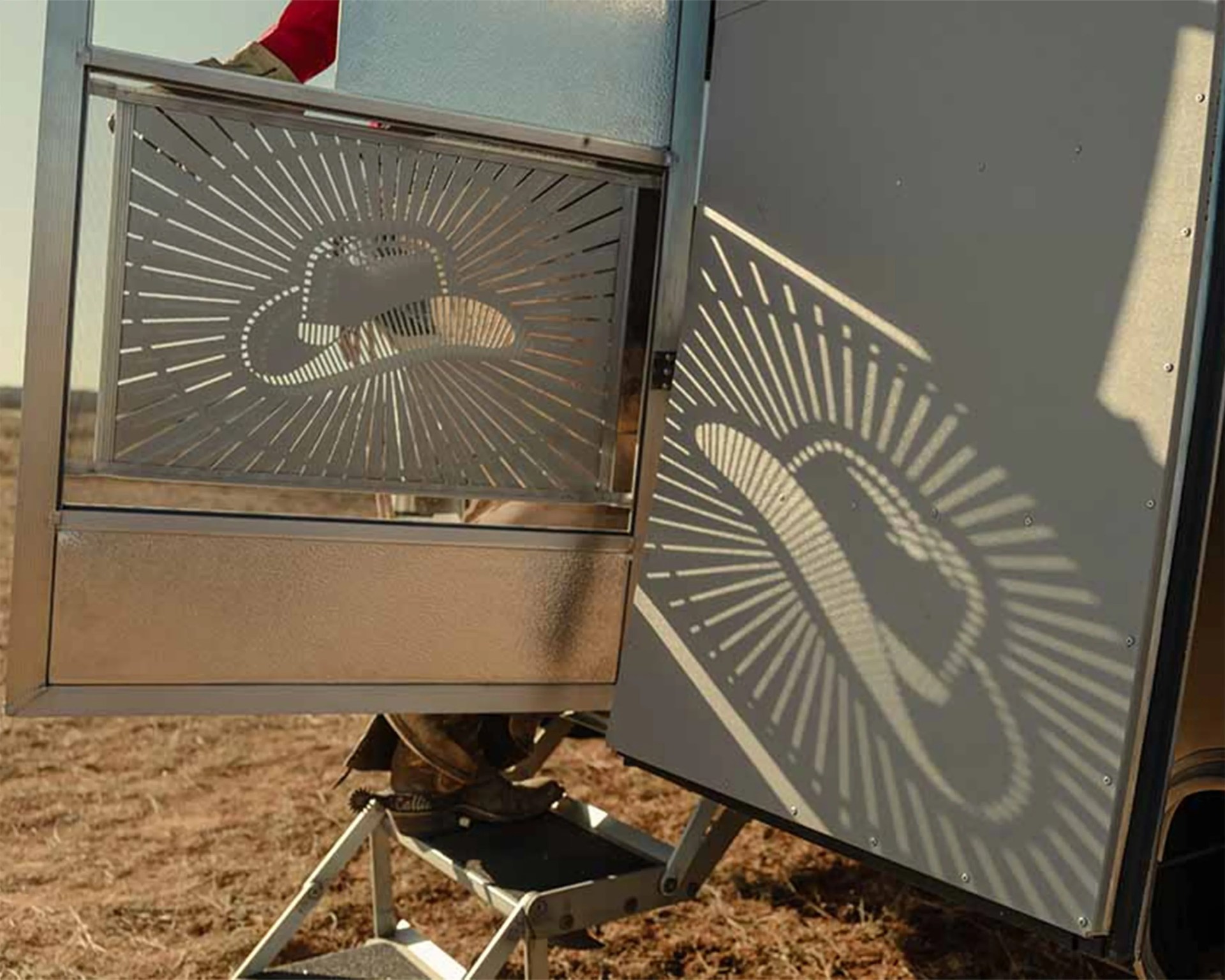Metal gate with a cutout cowboy hat design casting a shadow on a nearby surface, with a person wearing boots stepping on a metal step.