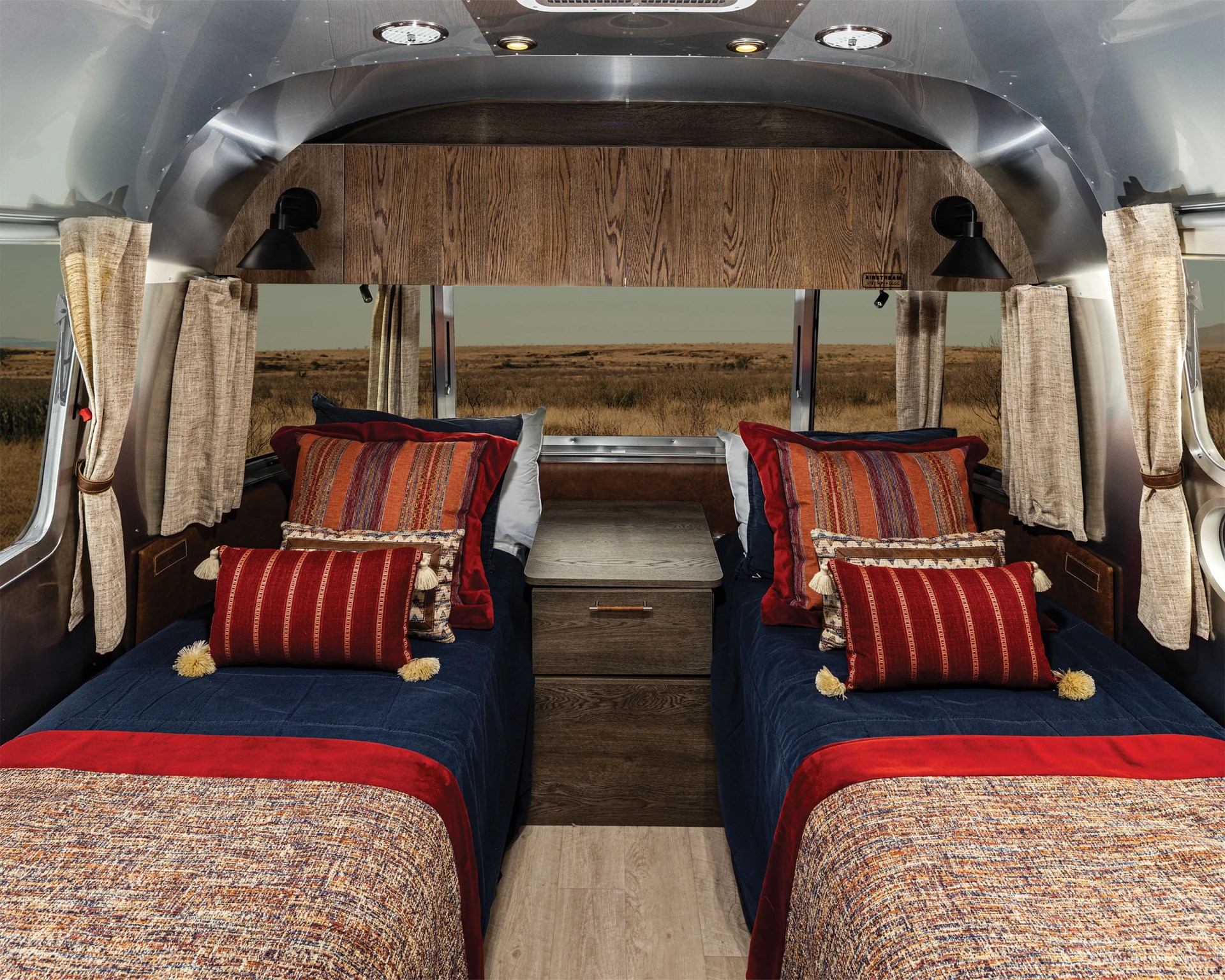 Interior of a camper with two twin beds featuring navy blue and red bedding, patterned pillows, beige curtains, and a wooden nightstand in the center.