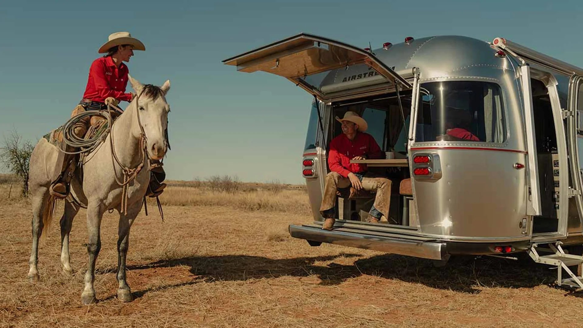 Person in a red shirt and cowboy hat riding a white horse near a silver Airstream trailer with an open hatch.