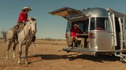 Person in a red shirt and cowboy hat riding a white horse near a silver Airstream trailer with an open hatch.