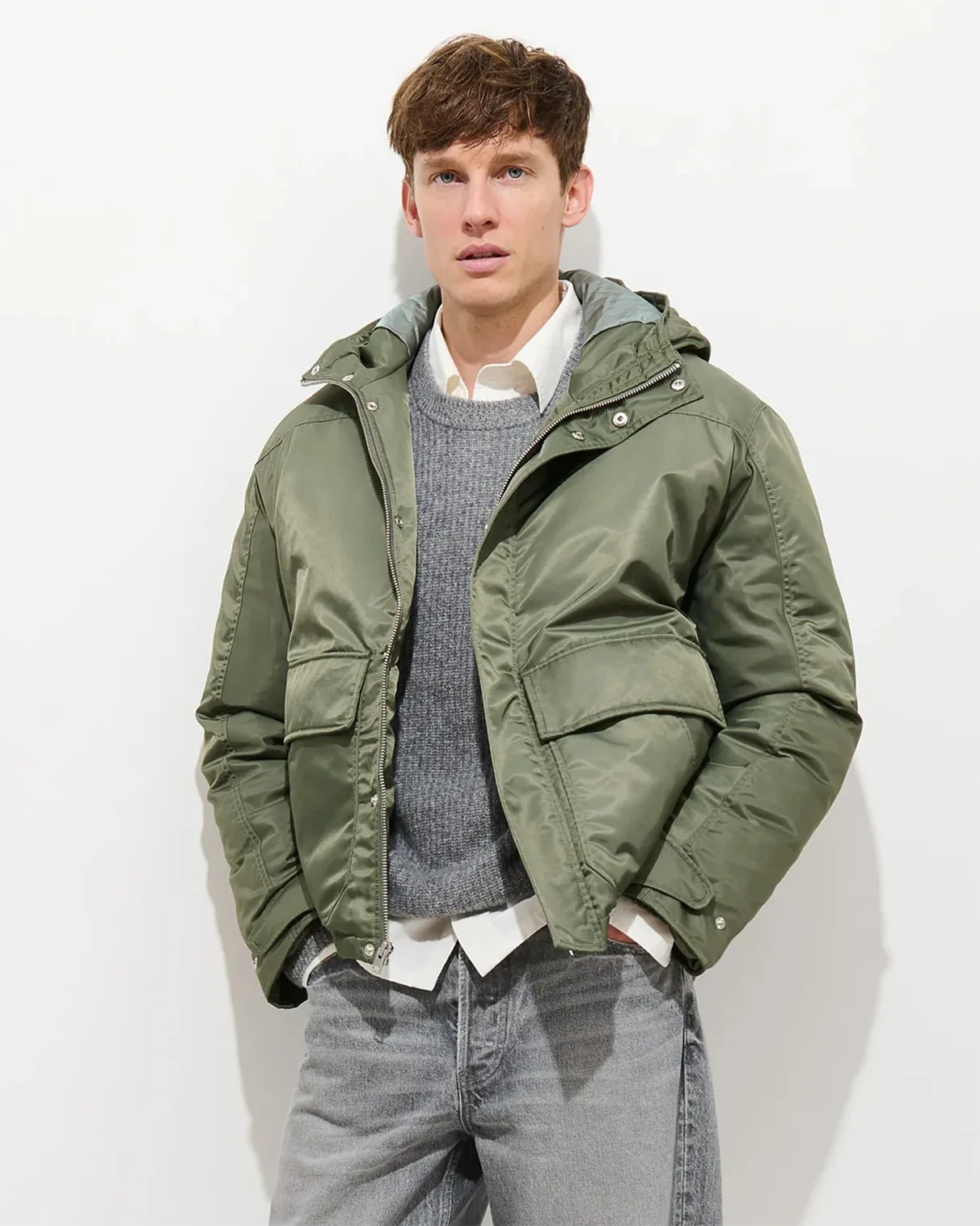 Man wearing an olive green hooded puffer jacket over a gray sweater and white shirt with gray jeans.