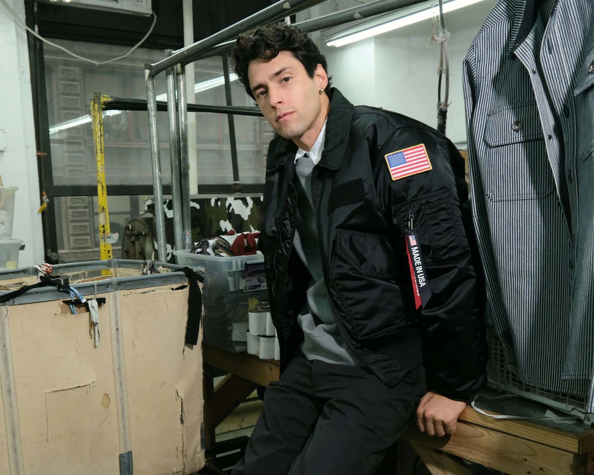 Man wearing a black bomber jacket with an American flag patch and "Made in USA" tag sitting on a wooden bench.