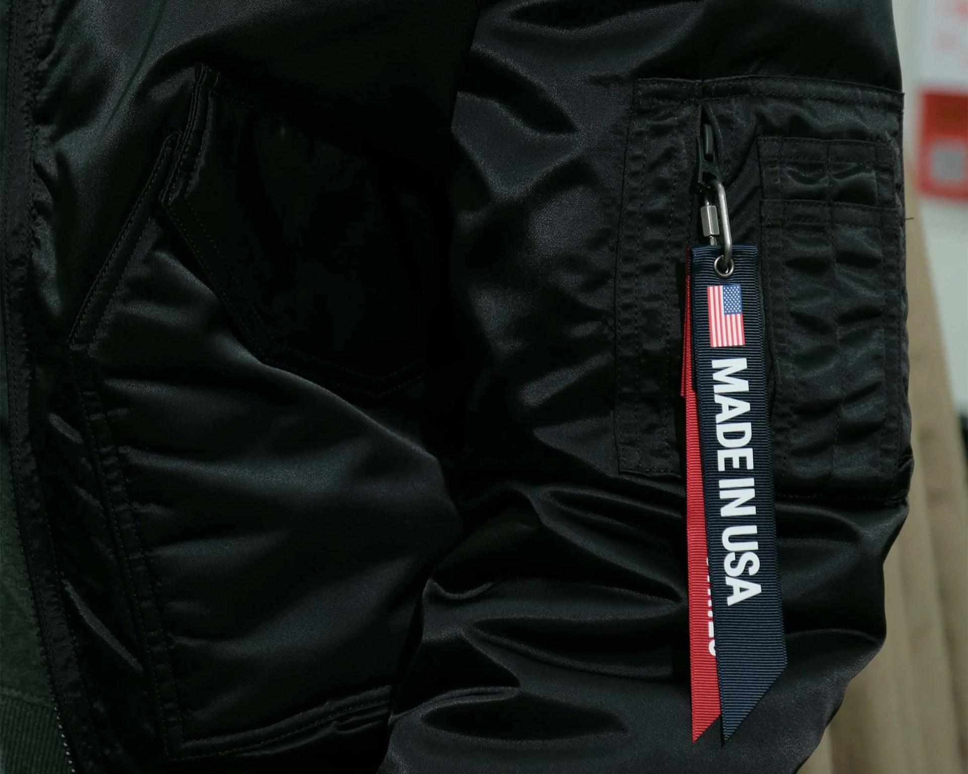 Black jacket sleeve with a zippered pocket and a "Made in USA" fabric tag attached by a metal clip.