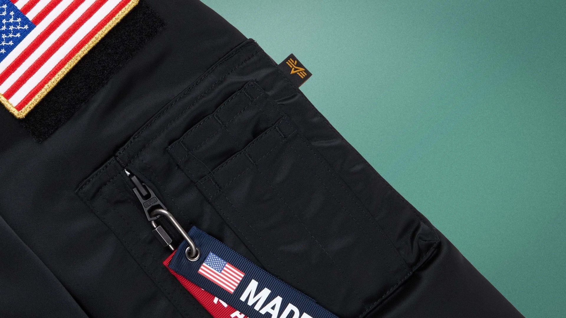 Black jacket sleeve with a zippered pocket, an American flag patch, and a "Made in USA" fabric tag attached to the zipper.