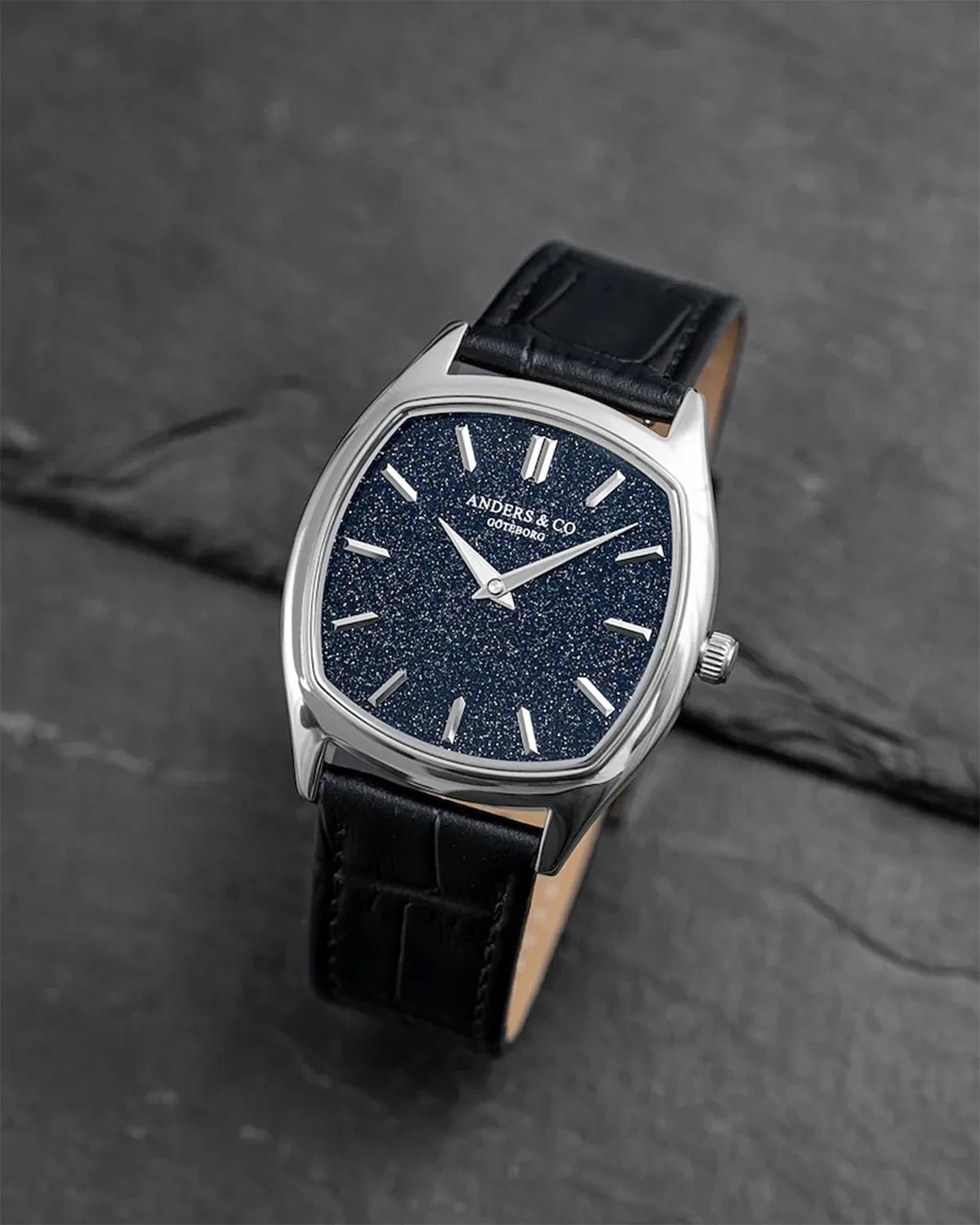 Silver-toned square watch with a glittery dark blue dial and black leather strap on a dark stone surface.