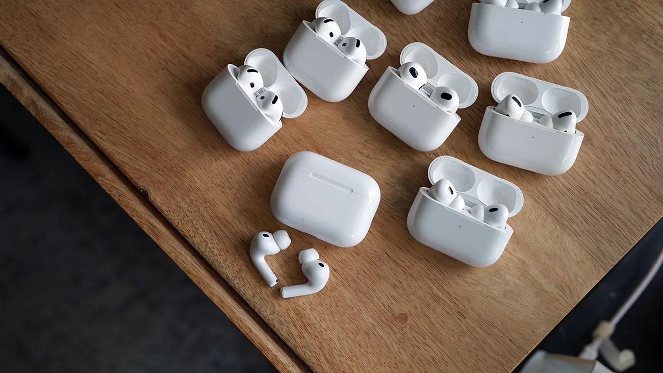 Seven pairs of white wireless earbuds in open charging cases and one pair outside the case on a wooden surface.