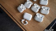 Seven pairs of white wireless earbuds in open charging cases and one pair outside the case on a wooden surface.