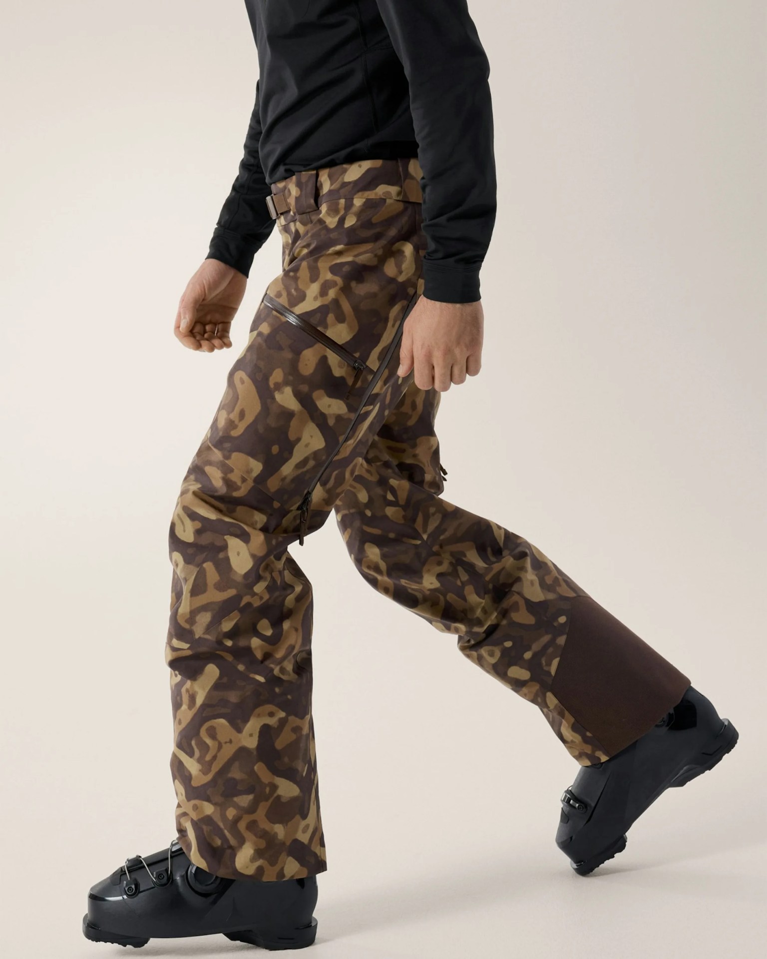 Brown and tan camouflage ski pants with zippered thigh pocket and reinforced dark brown cuffs worn with black ski boots.