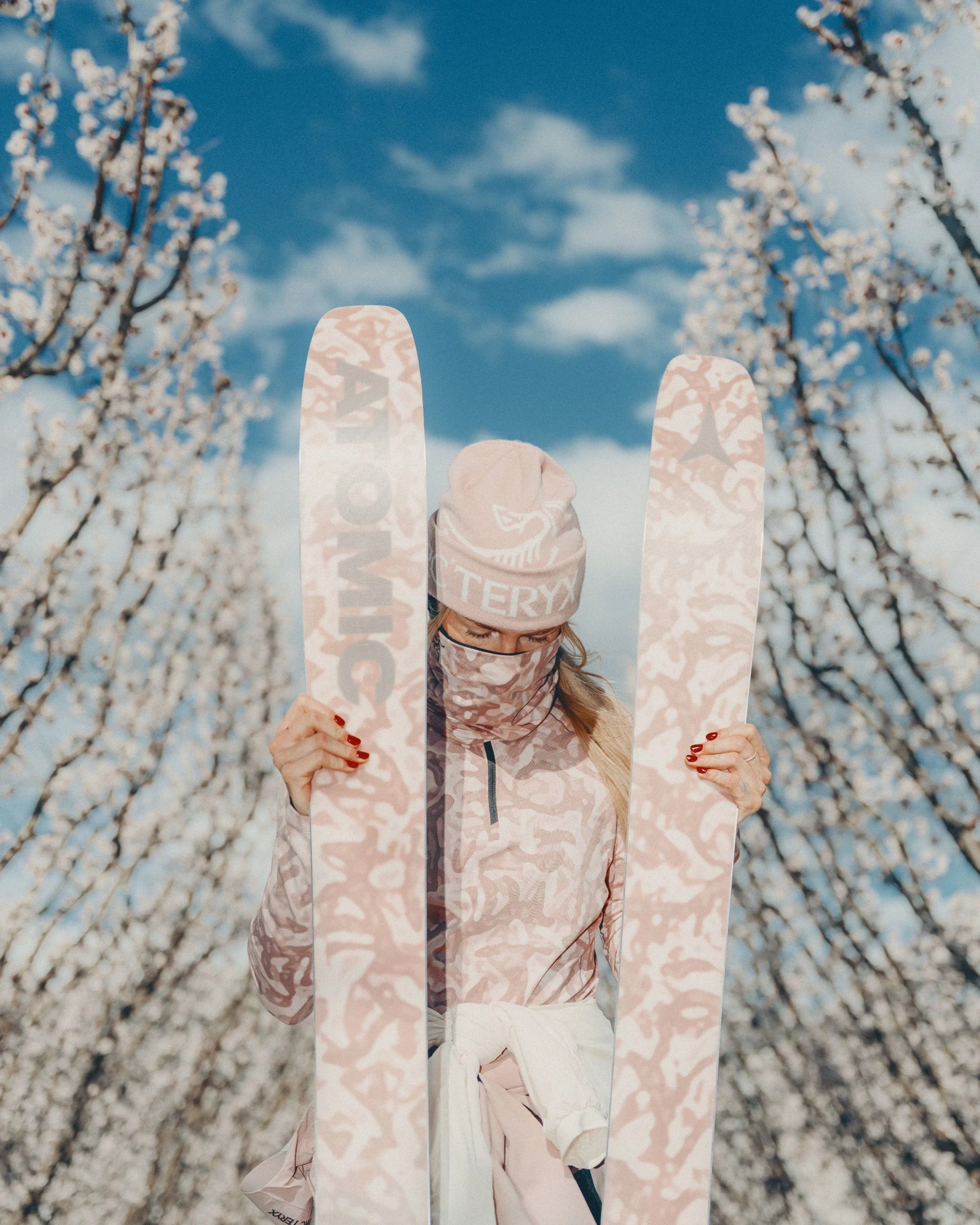 Person wearing pink patterned ski outfit and hat holding matching Atomic skis in front of blossoming trees.