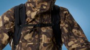 Brown and tan camouflage jacket with black backpack straps and a chest buckle against a blue background.