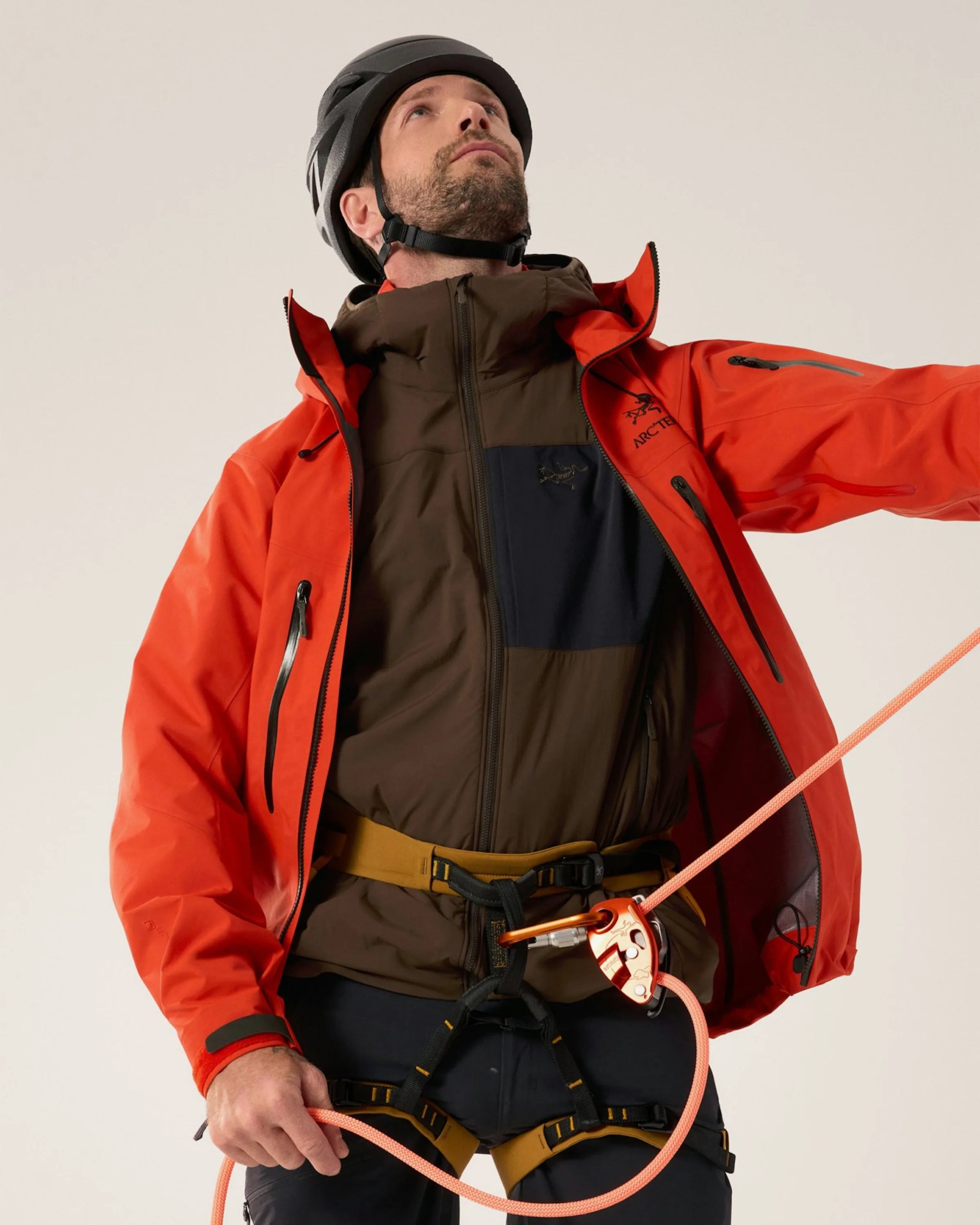 Man wearing a black helmet, red Arc'teryx jacket, brown jacket, and climbing harness holding an orange climbing rope.